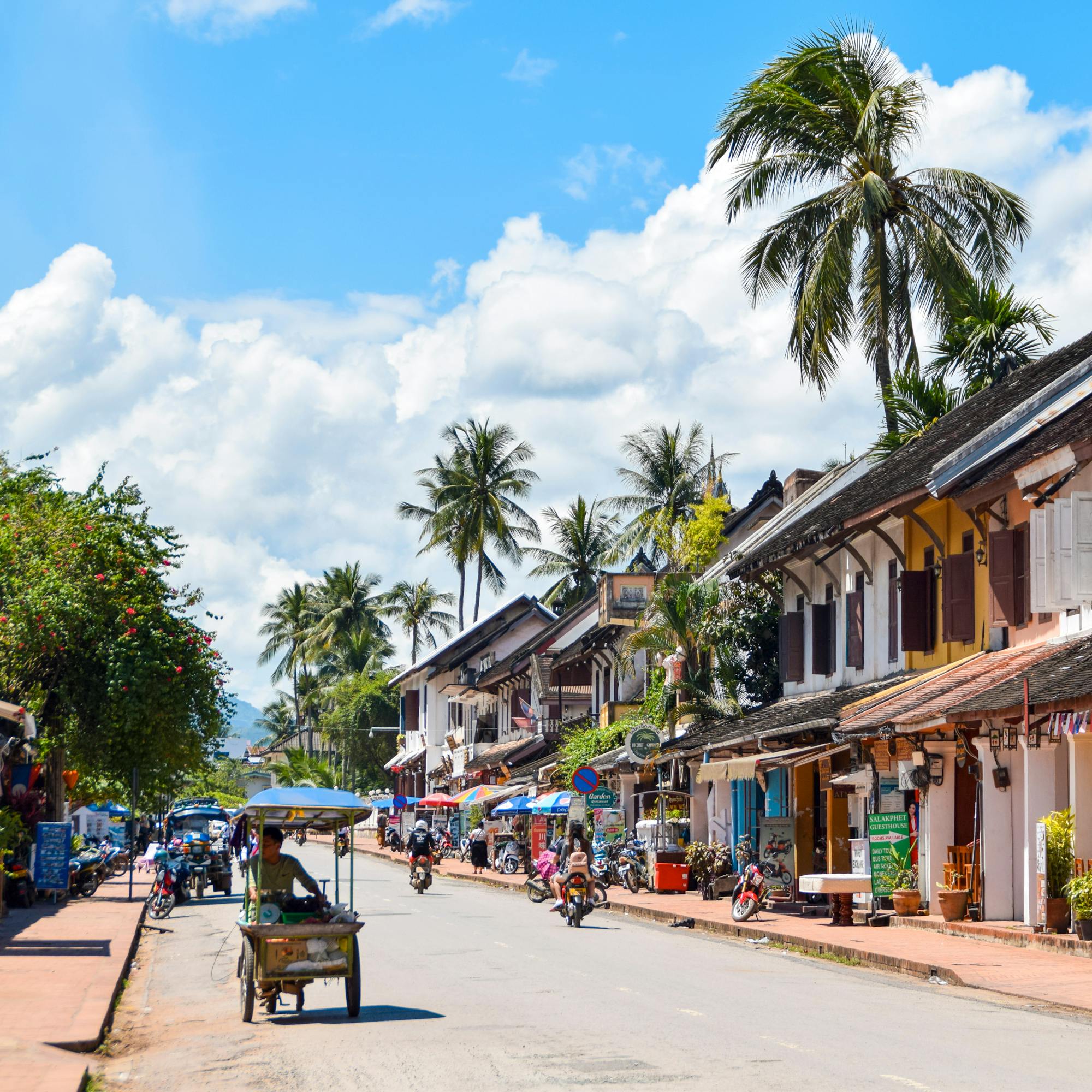 Sunlit street lined with low buildings and palm trees, with scooters and pedestrians in the distance under soft daylight.