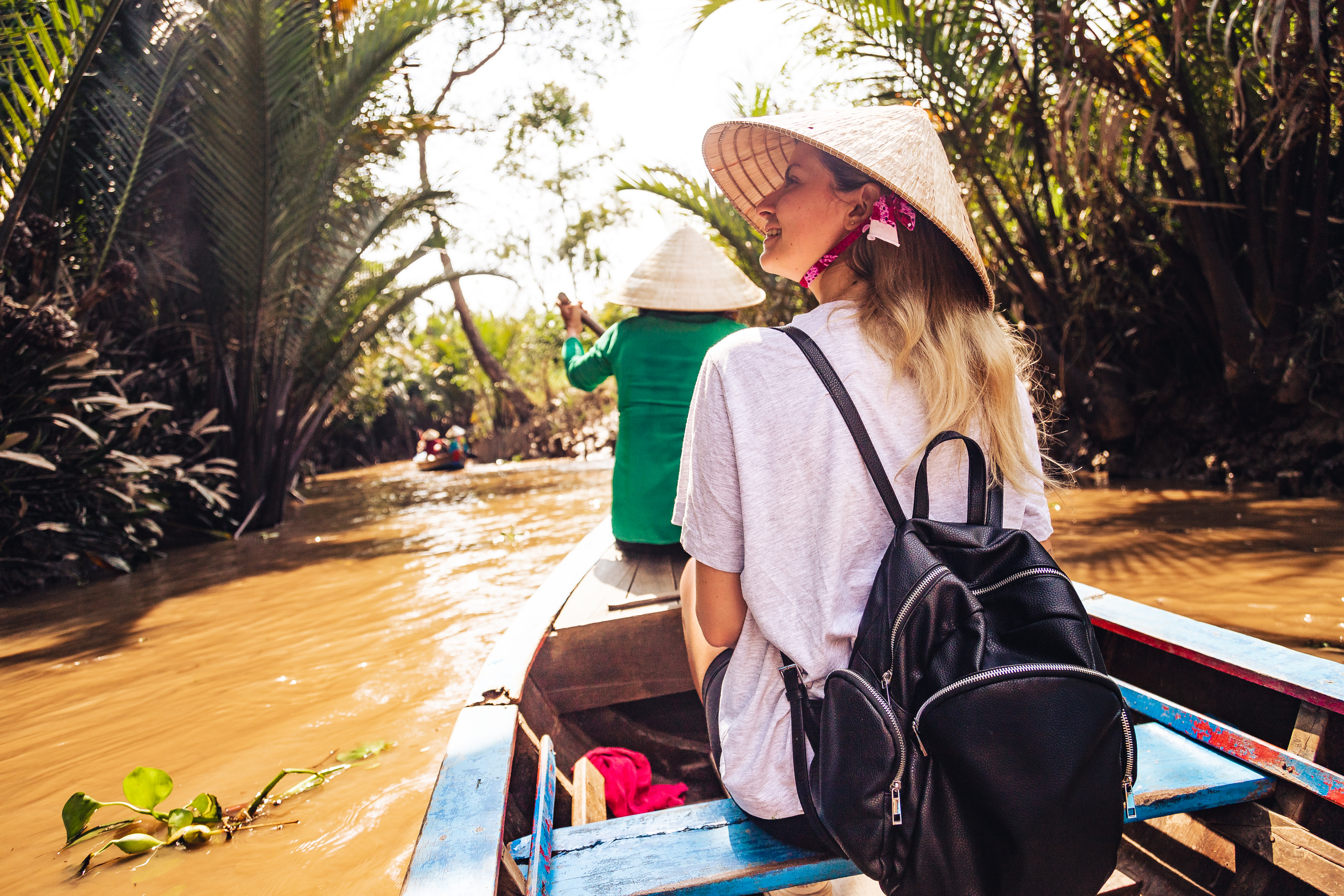 Person rides in a longtail boat along a narrow canal, with dense palms and muddy water stretching ahead under soft daylight.
