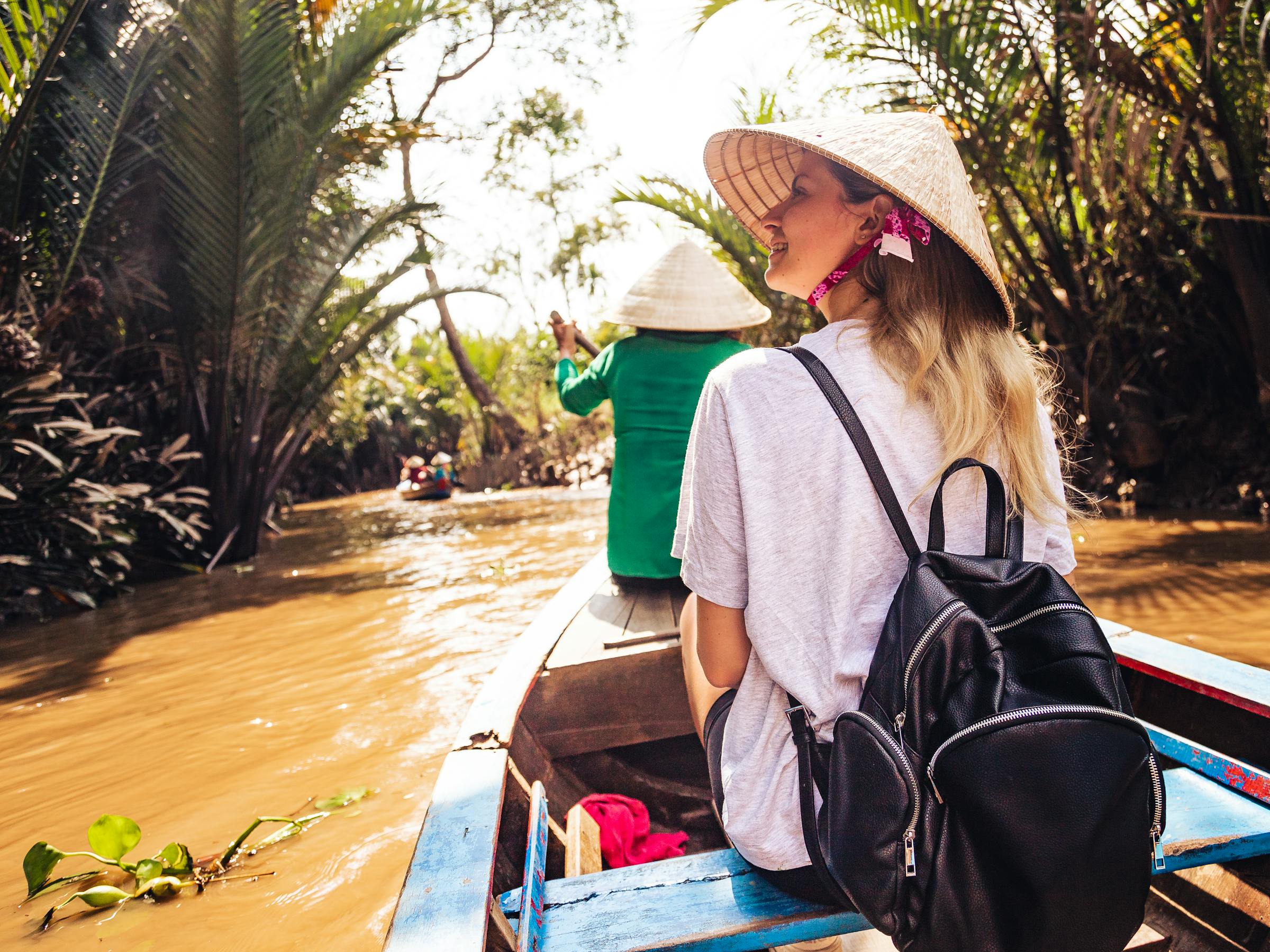 Person rides in a longtail boat along a narrow canal, with dense palms and muddy water stretching ahead under soft daylight.