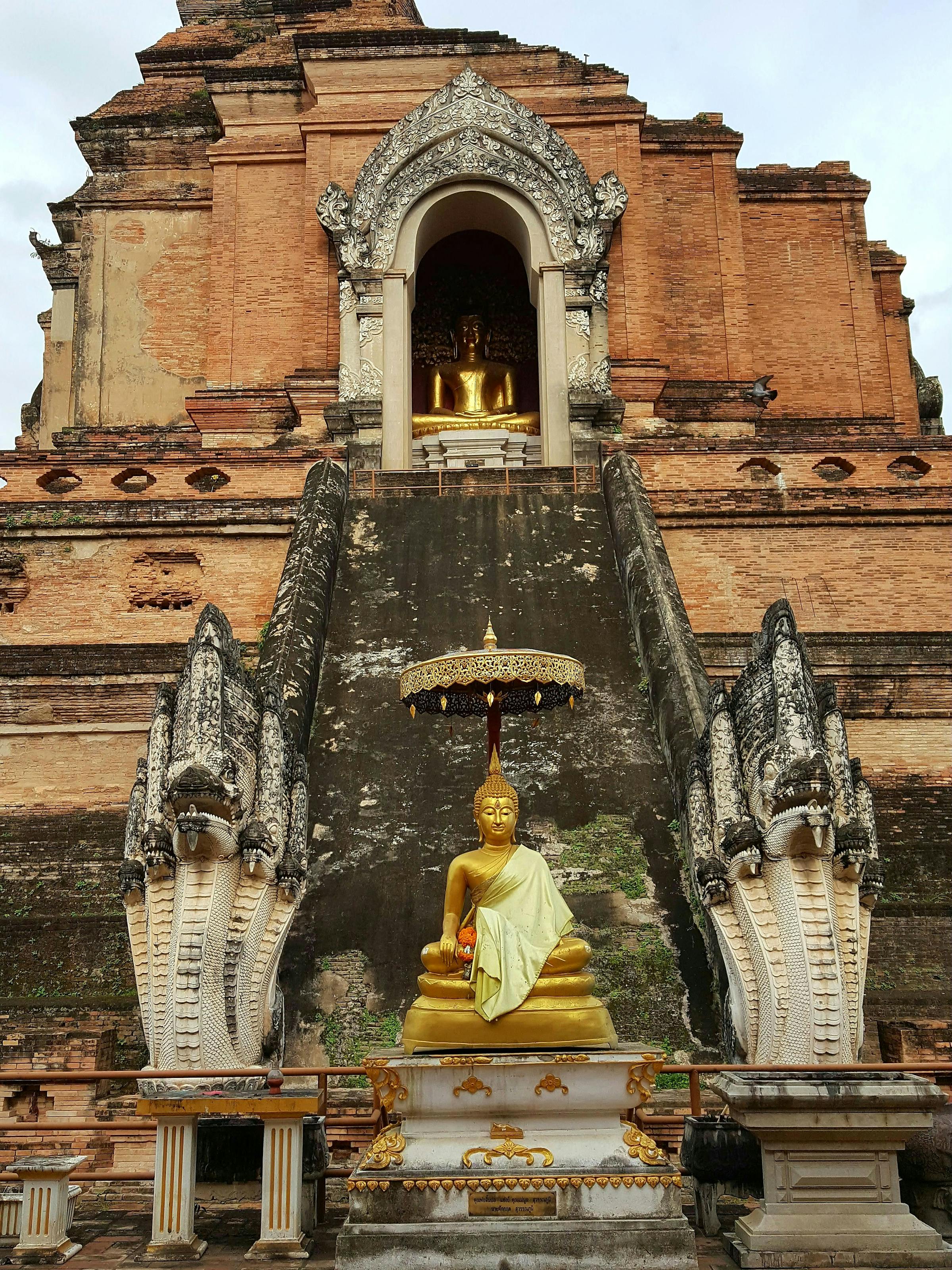 Ancient temple ruins with carved stone walls and a weathered arch, surrounded by trees and cloudy sky under soft daylight.