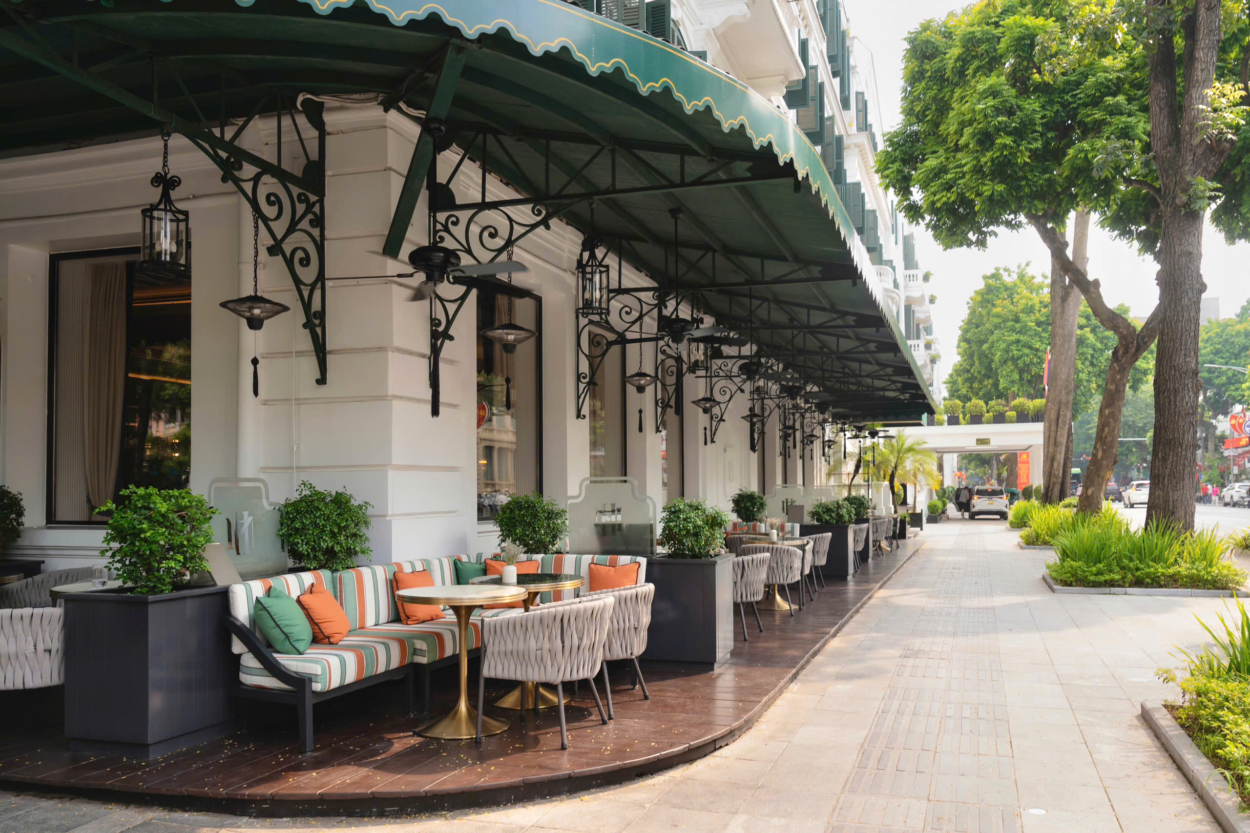 Outdoor terrace lined with tables and chairs beneath green awnings, with potted plants and arched windows.