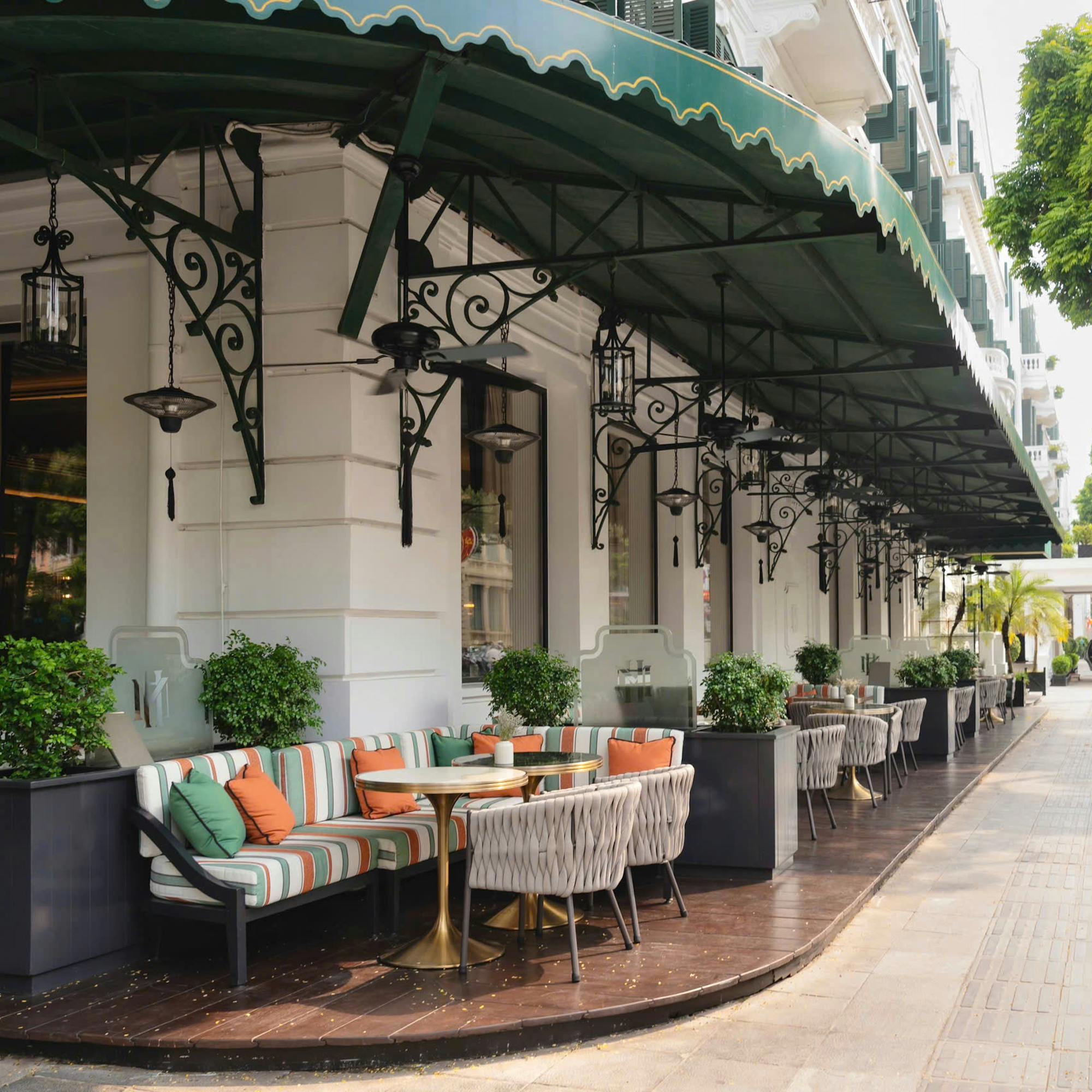 Outdoor terrace lined with tables and chairs beneath green awnings, with potted plants and arched windows.
