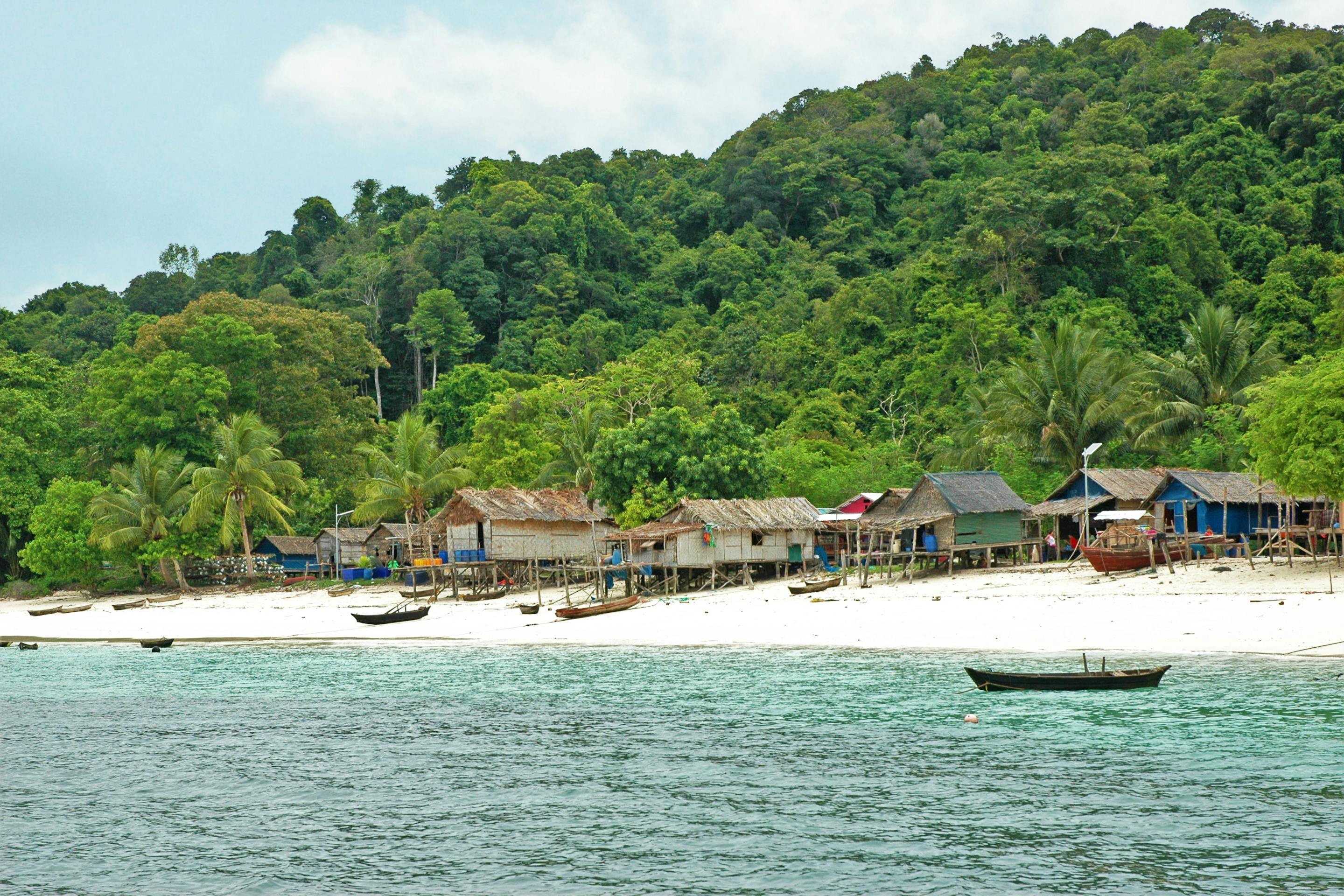 Small seaside village lines a white-sand beach, with jungle hills behind and shallow turquoise water under soft daylight.