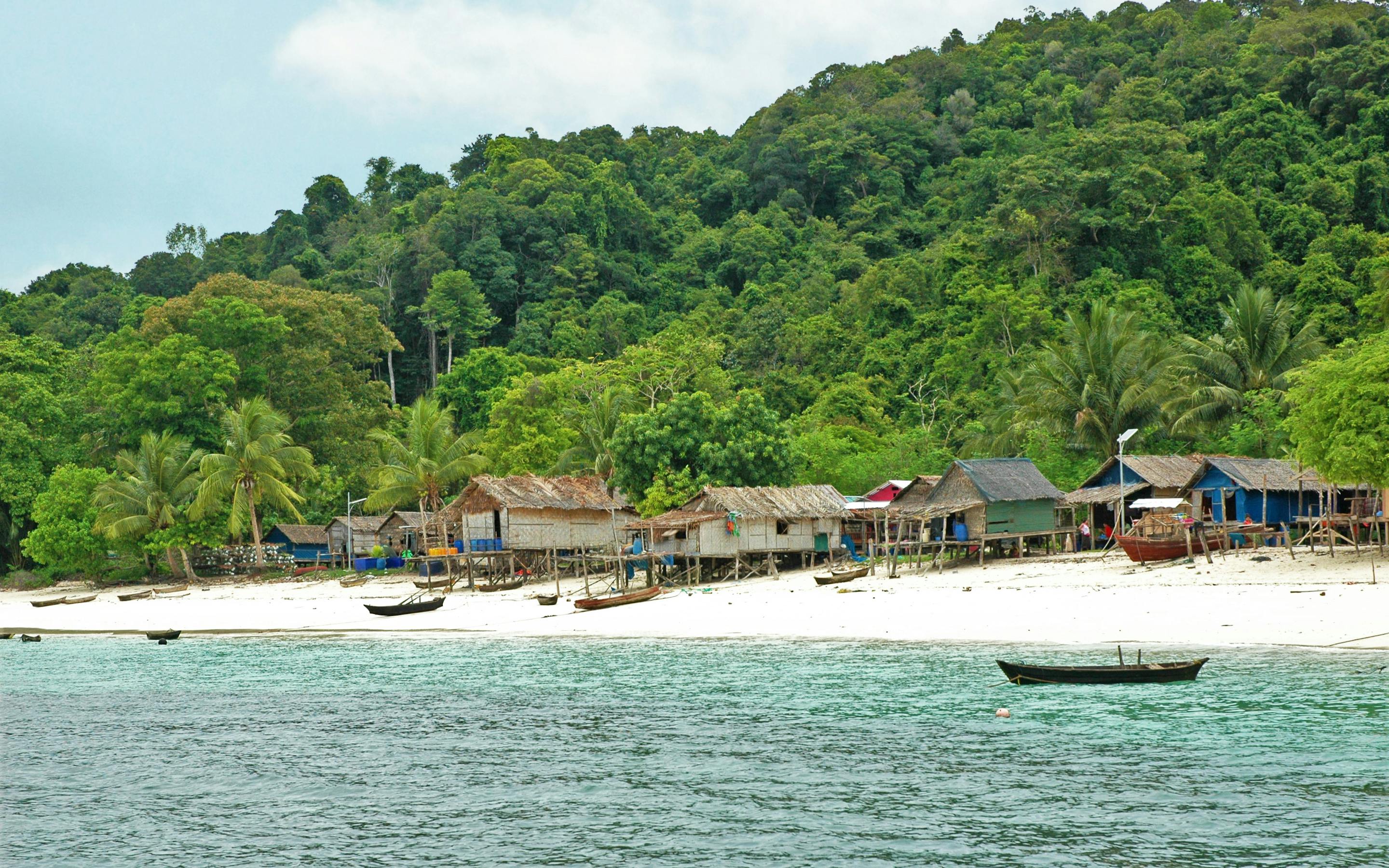 Small seaside village lines a white-sand beach, with jungle hills behind and shallow turquoise water under soft daylight.