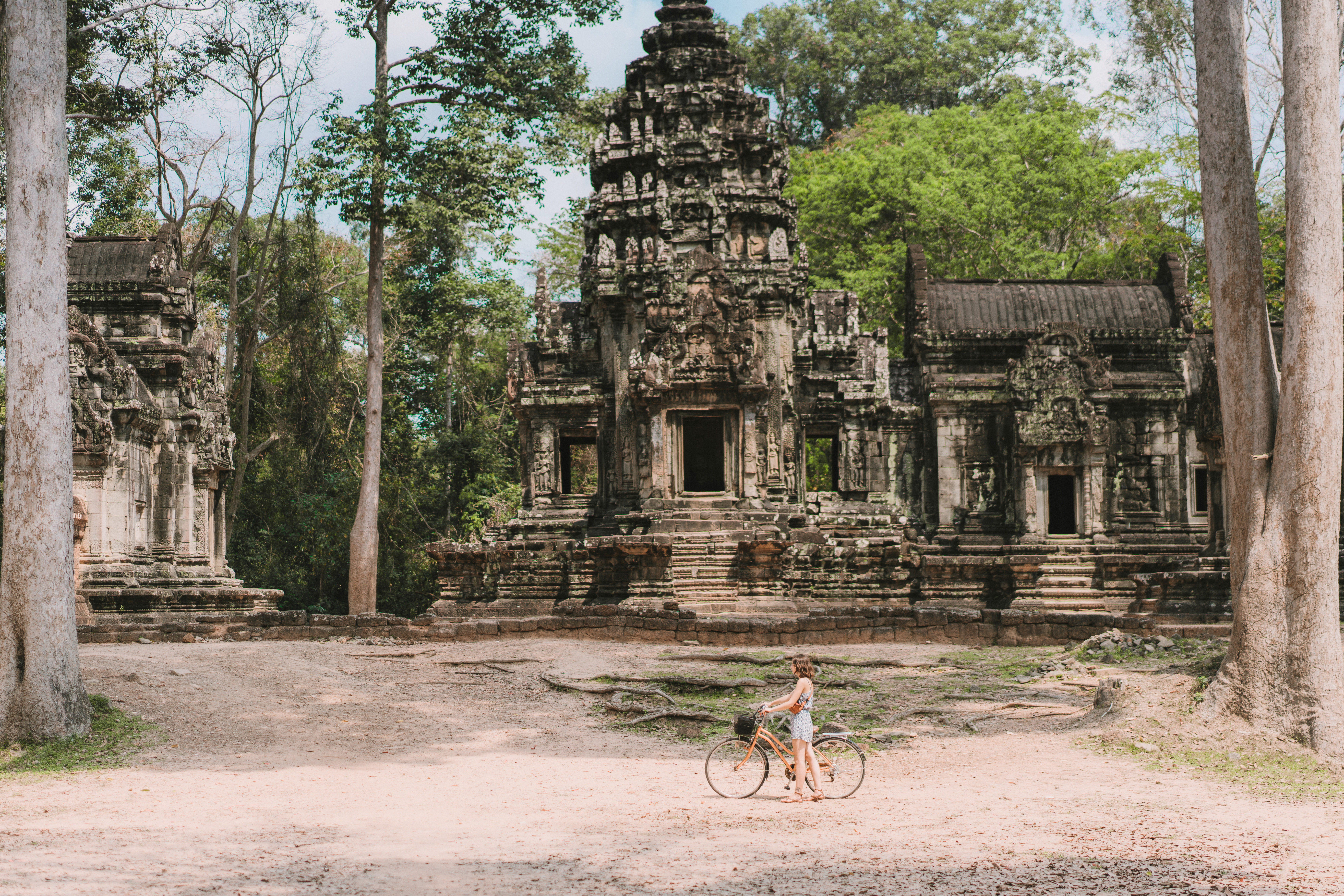 Cyclist rides past weathered stone temple ruins, with carved walls and trees surrounding the path.