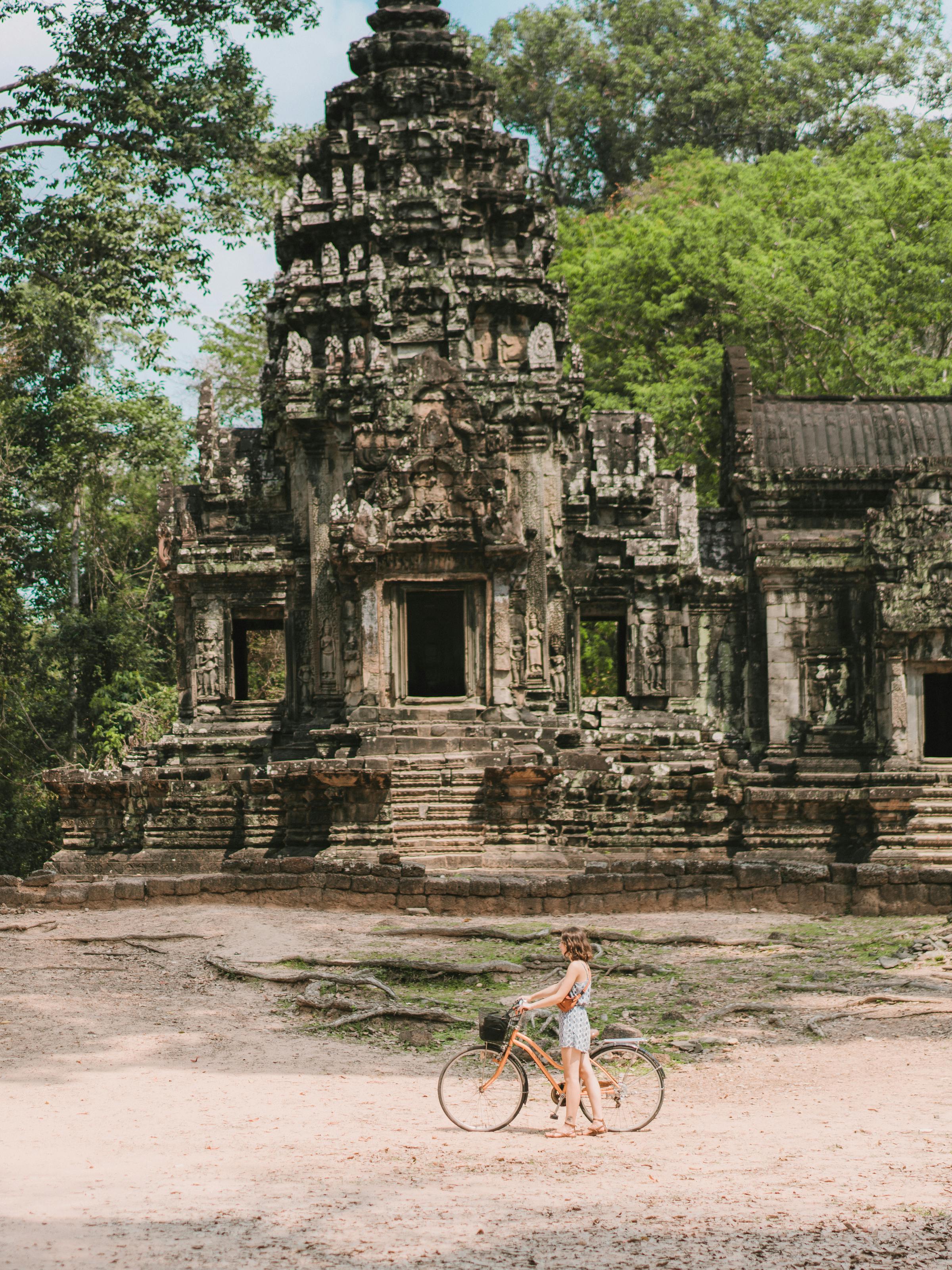 Cyclist rides past weathered stone temple ruins, with carved walls and trees surrounding the path.