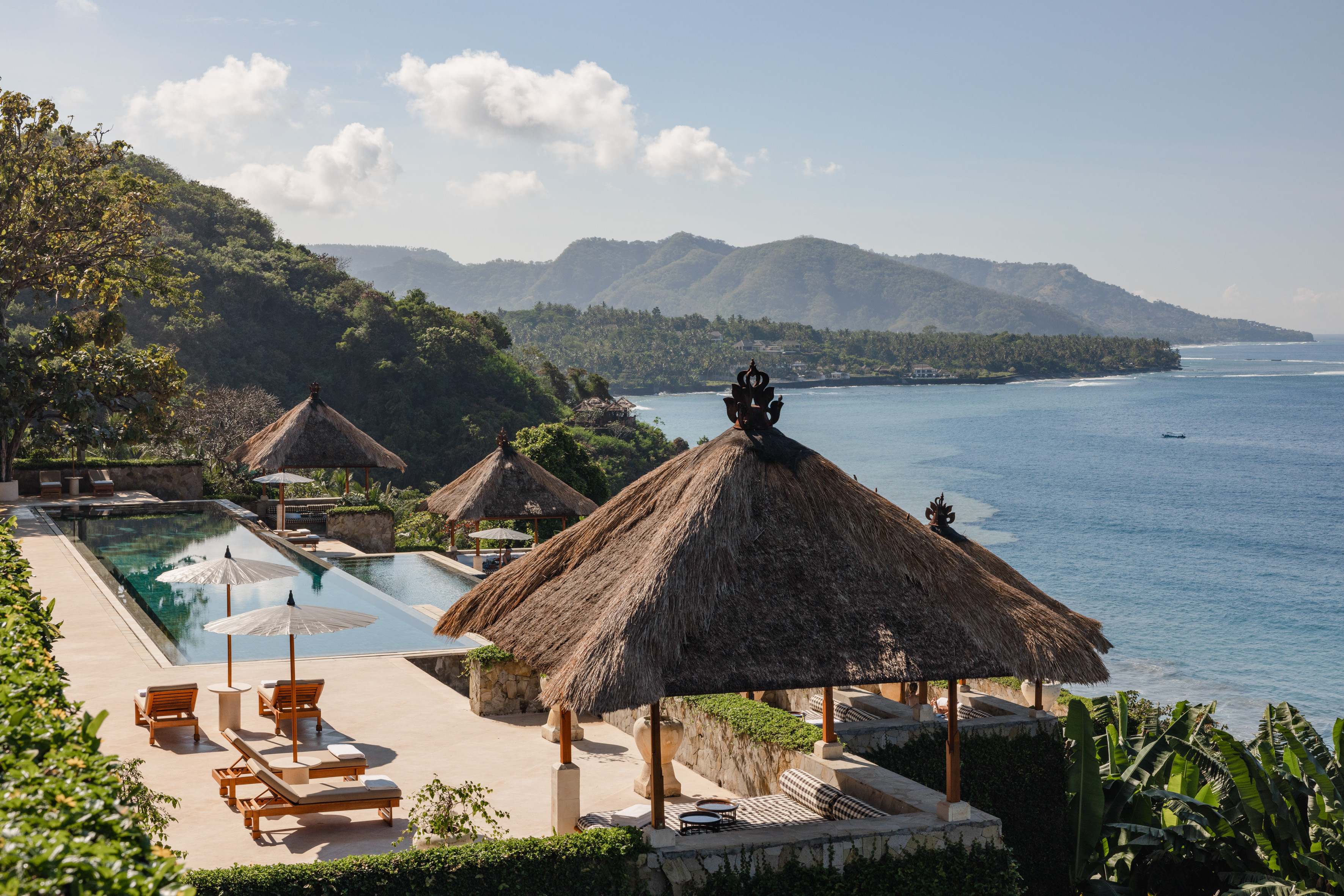 Thatched gazebo overlooks a pool and ocean view, with mountains in background under soft daylight.
