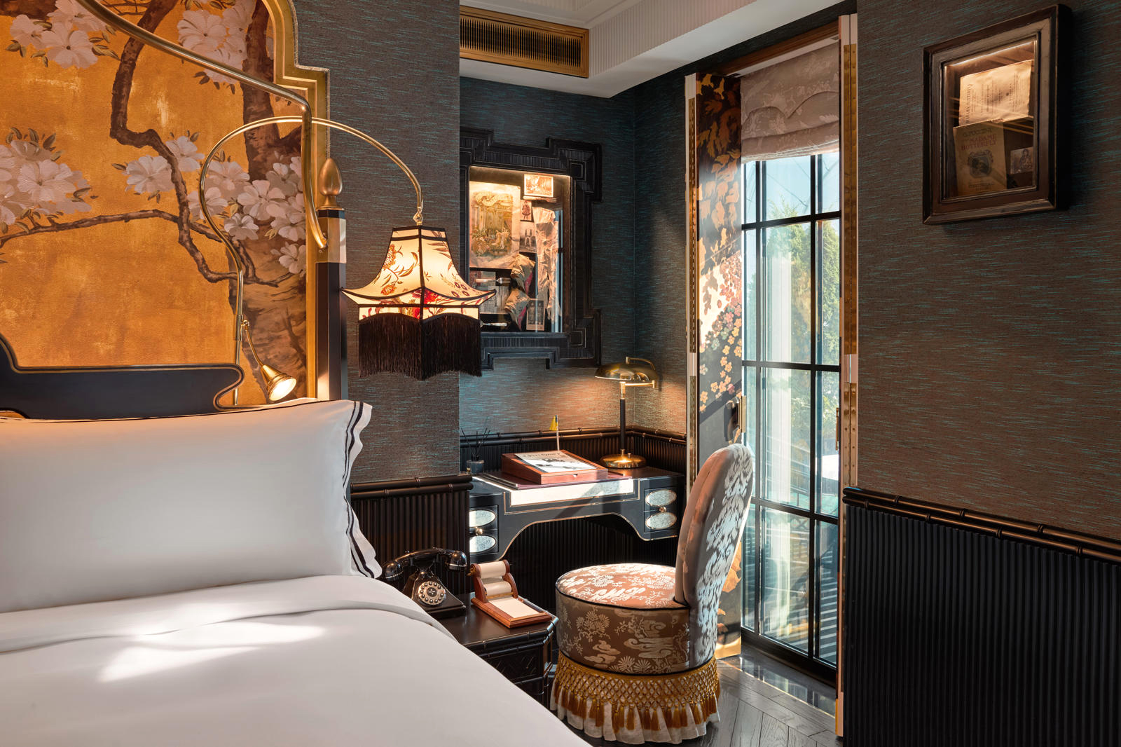 Hotel bedroom with a neatly made king bed, bedside lamps, and framed art beside a windowed sitting area under soft daylight.