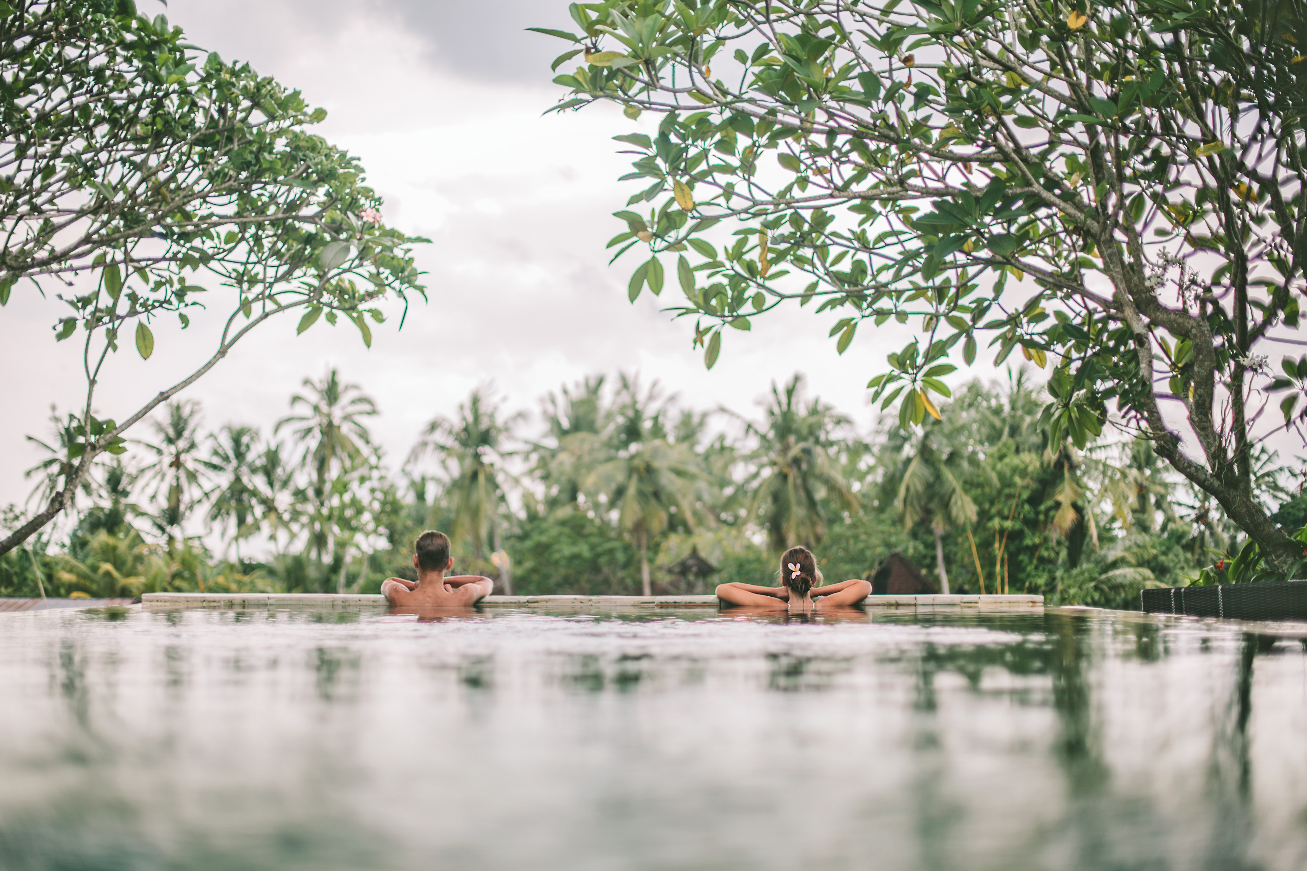 Two swimmers bathe in calm waters among riverside trees,  and reflections on the surface.
