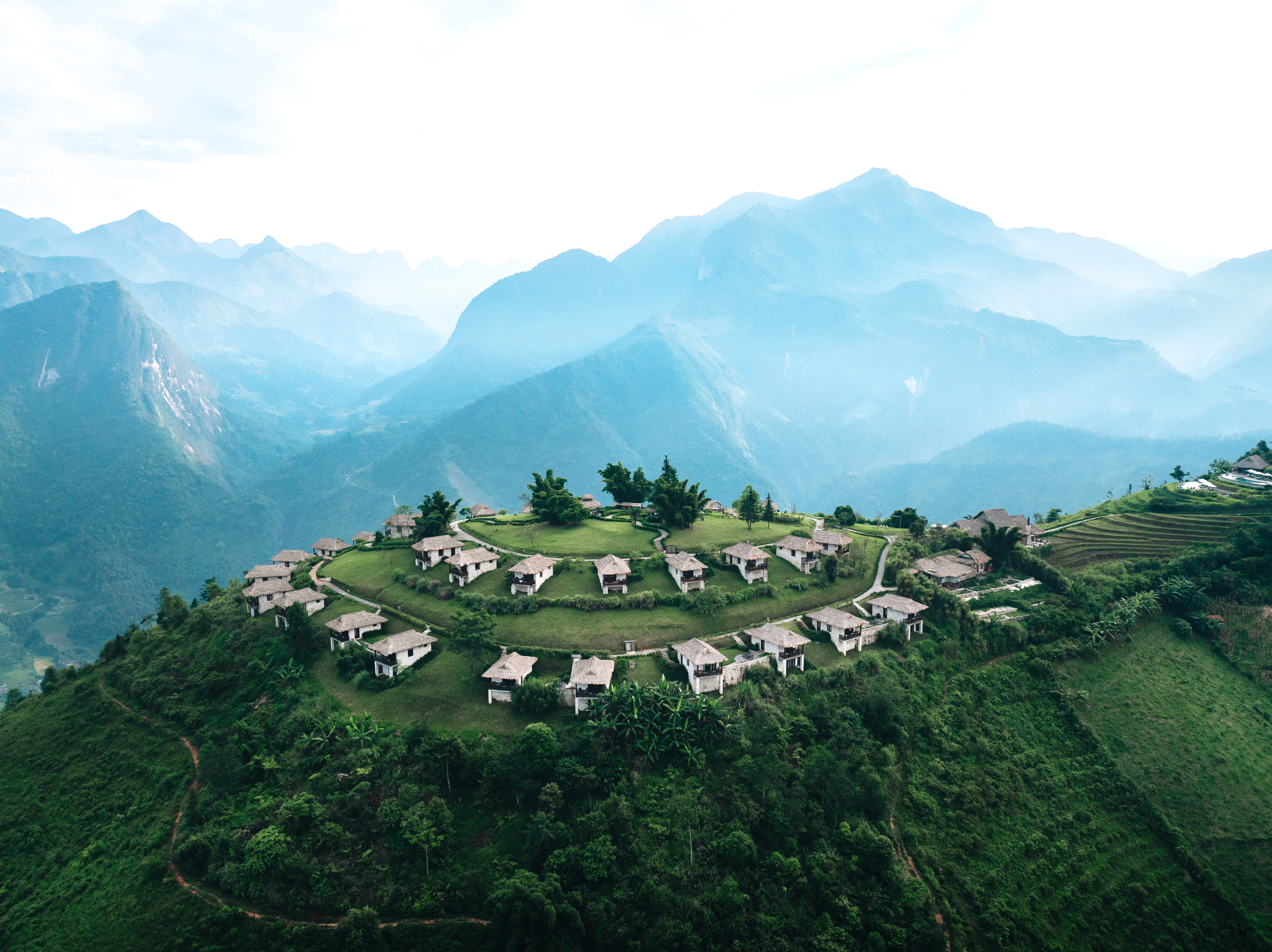 Aerial view of hillside lodges on a green ridge, with winding paths and blue mountains in the distance under soft daylight.