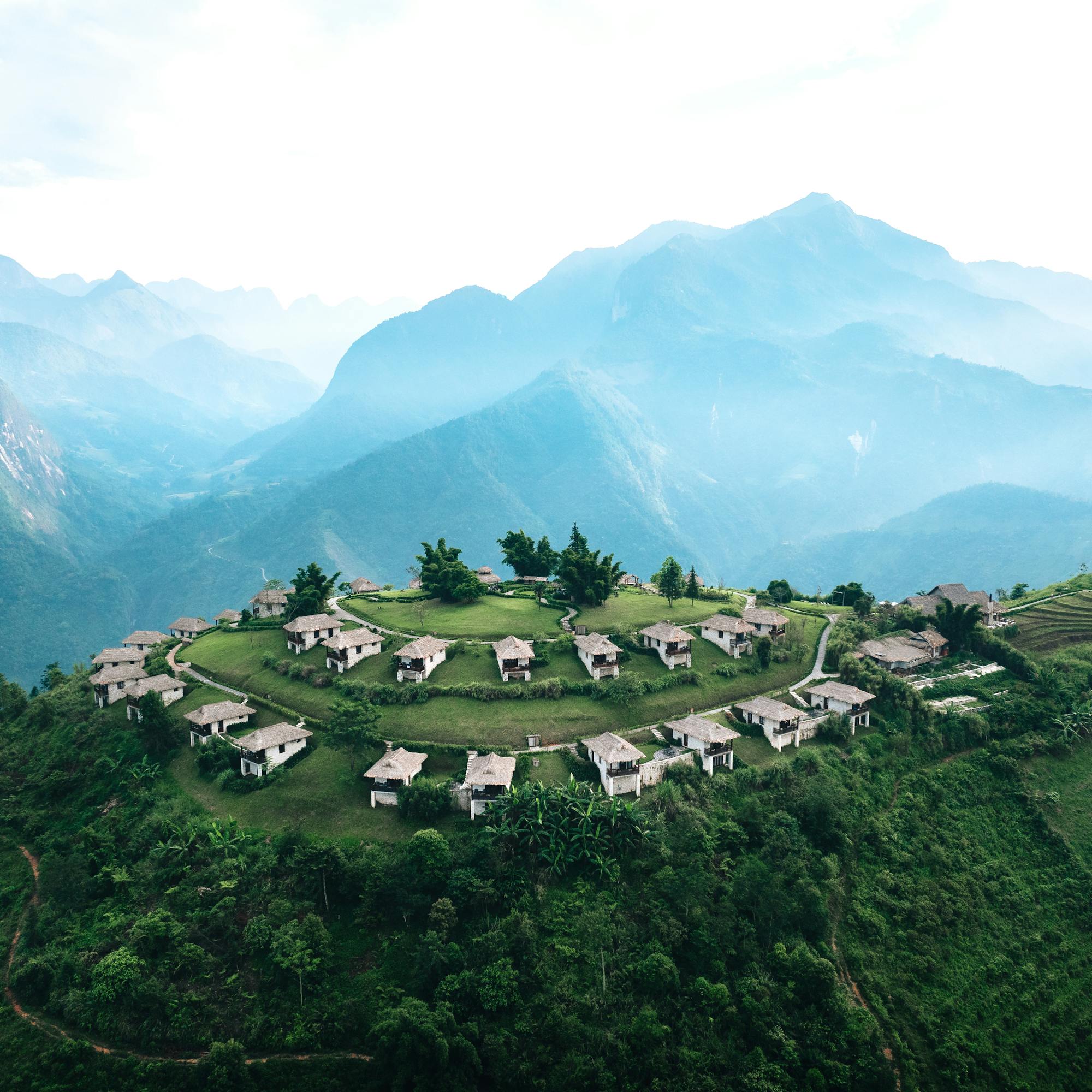 Aerial view of hillside lodges on a green ridge, with winding paths and blue mountains in the distance under soft daylight.