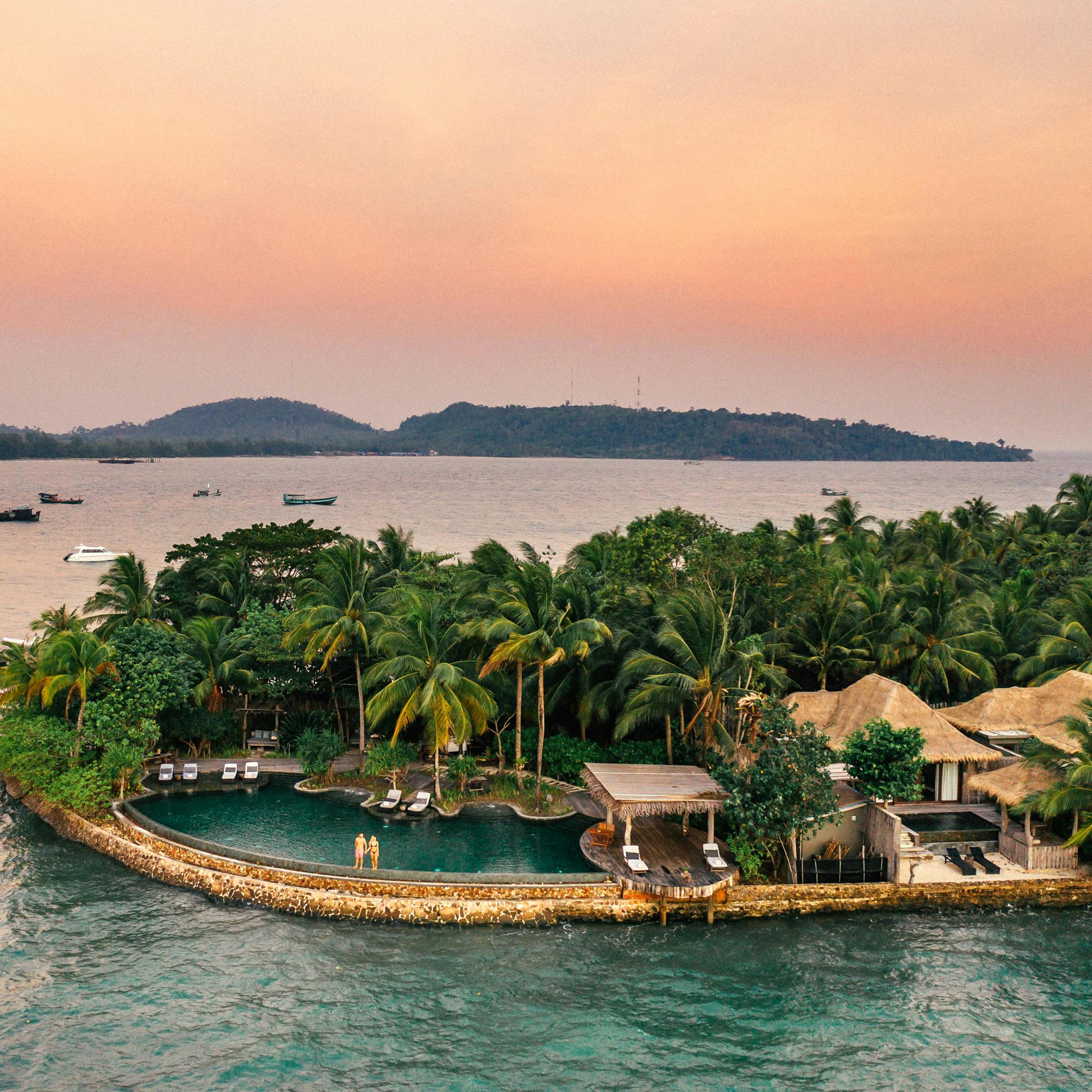 Aerial view of a small island resort with a pool, surrounded by turquoise water and dense palm trees under soft daylight.