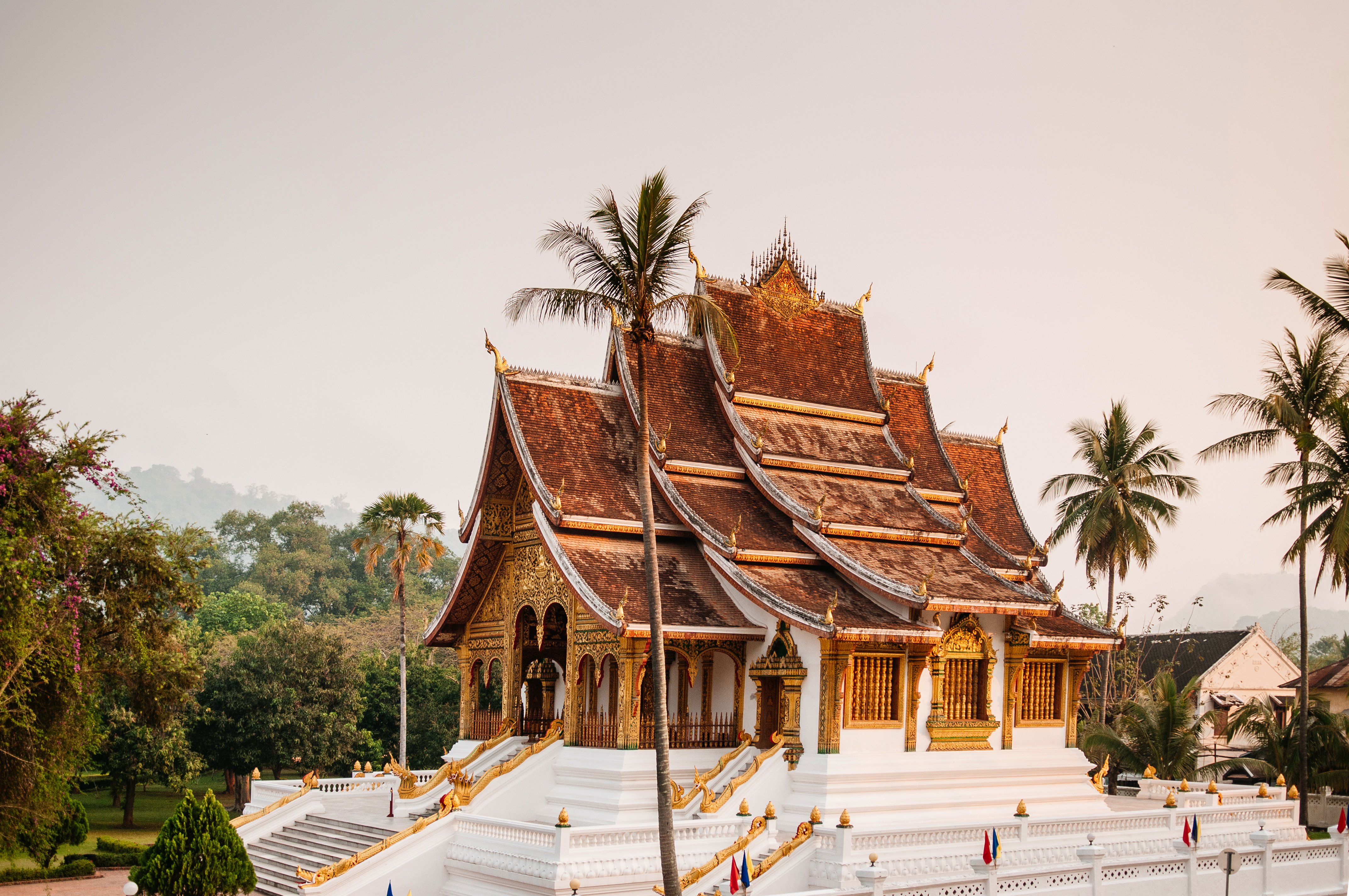 Ornate temple rooflines rise above a white terrace, with palm trees and a pale sky in the background under soft daylight.