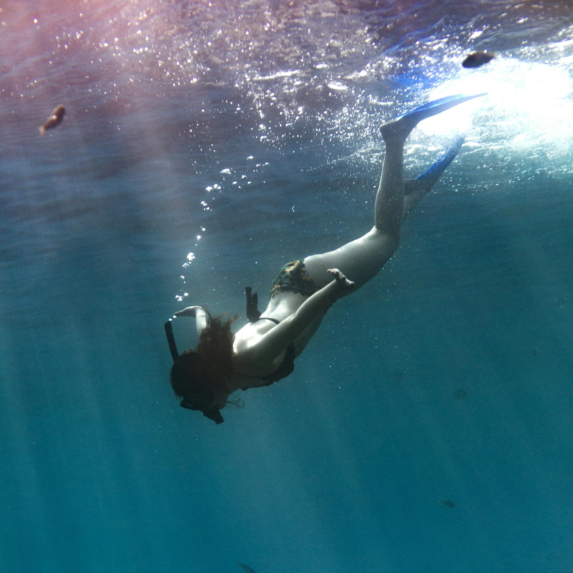 Snorkeler swims underwater near a reef wall, with bubbles rising and deep blue water in the background.