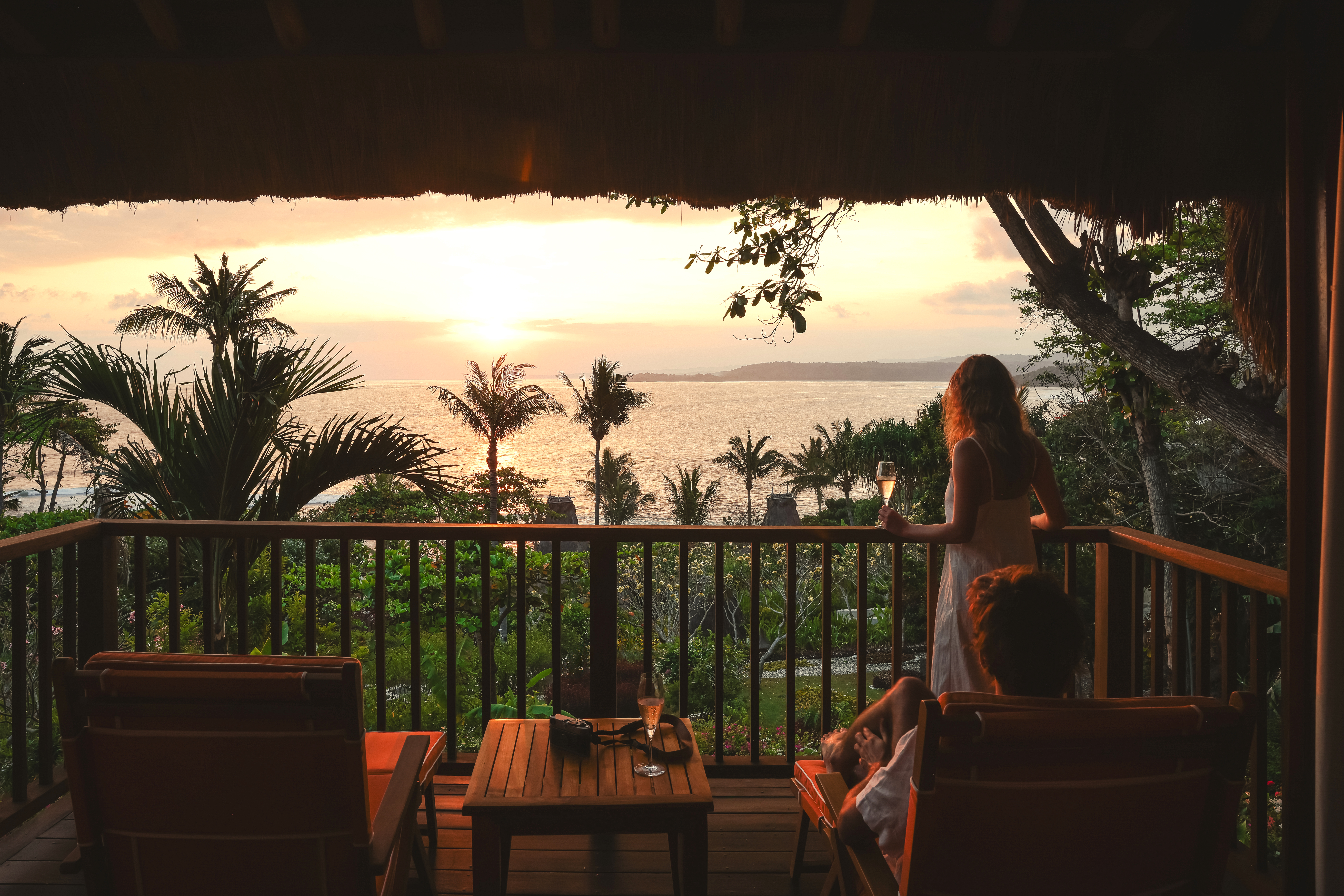 Person stands on a terrace facing a sunset over water, with palm fronds and a warm orange sky.