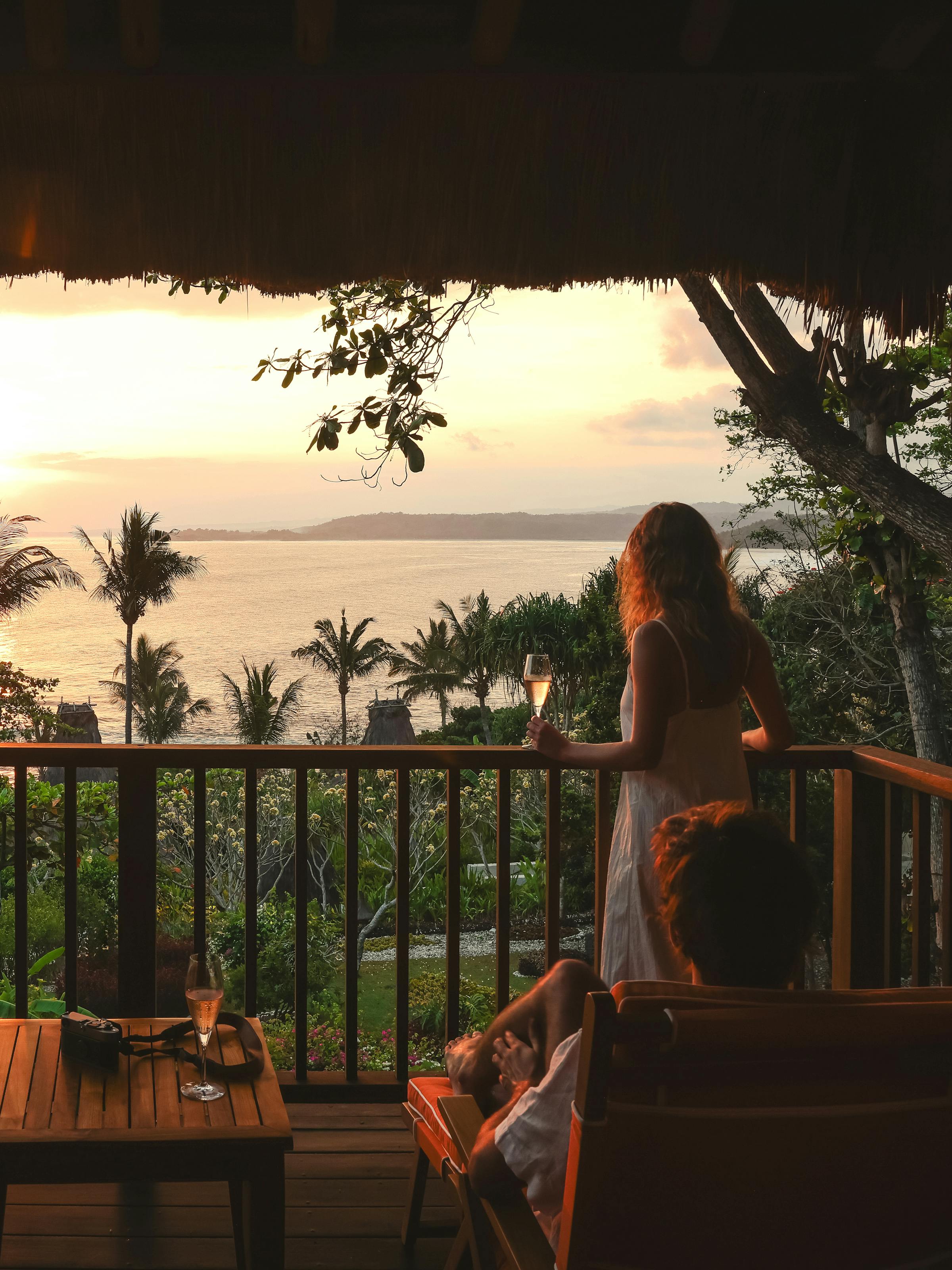 Person stands on a terrace facing a sunset over water, with palm fronds and a warm orange sky.