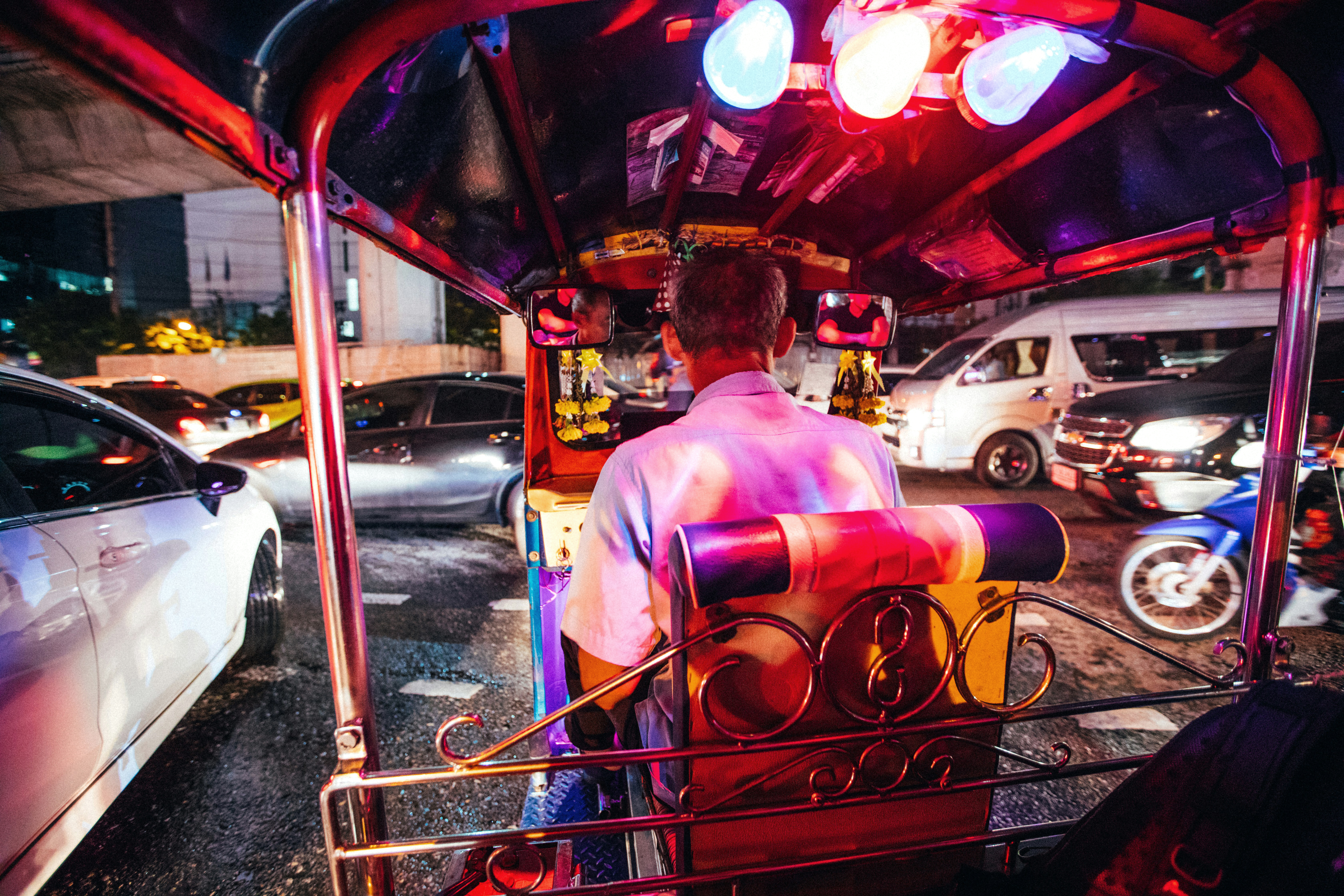 Tuk-tuk driver rides through a neon-lit street at night, with blurred headlights and colorful reflections.