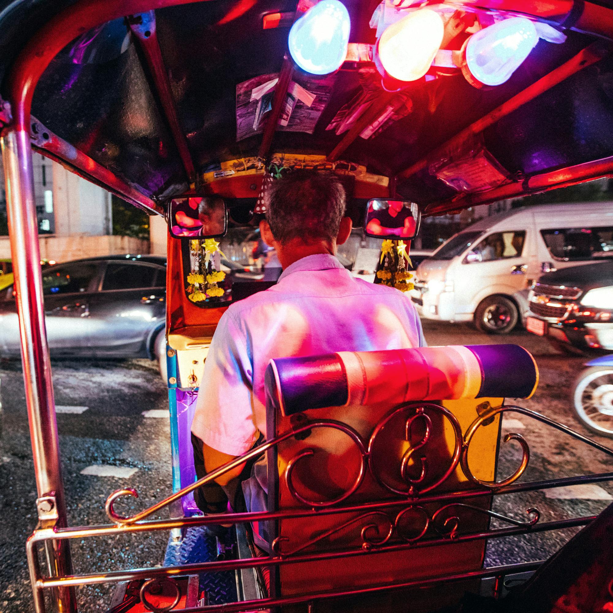 Tuk-tuk driver rides through a neon-lit street at night, with blurred headlights and colorful reflections.