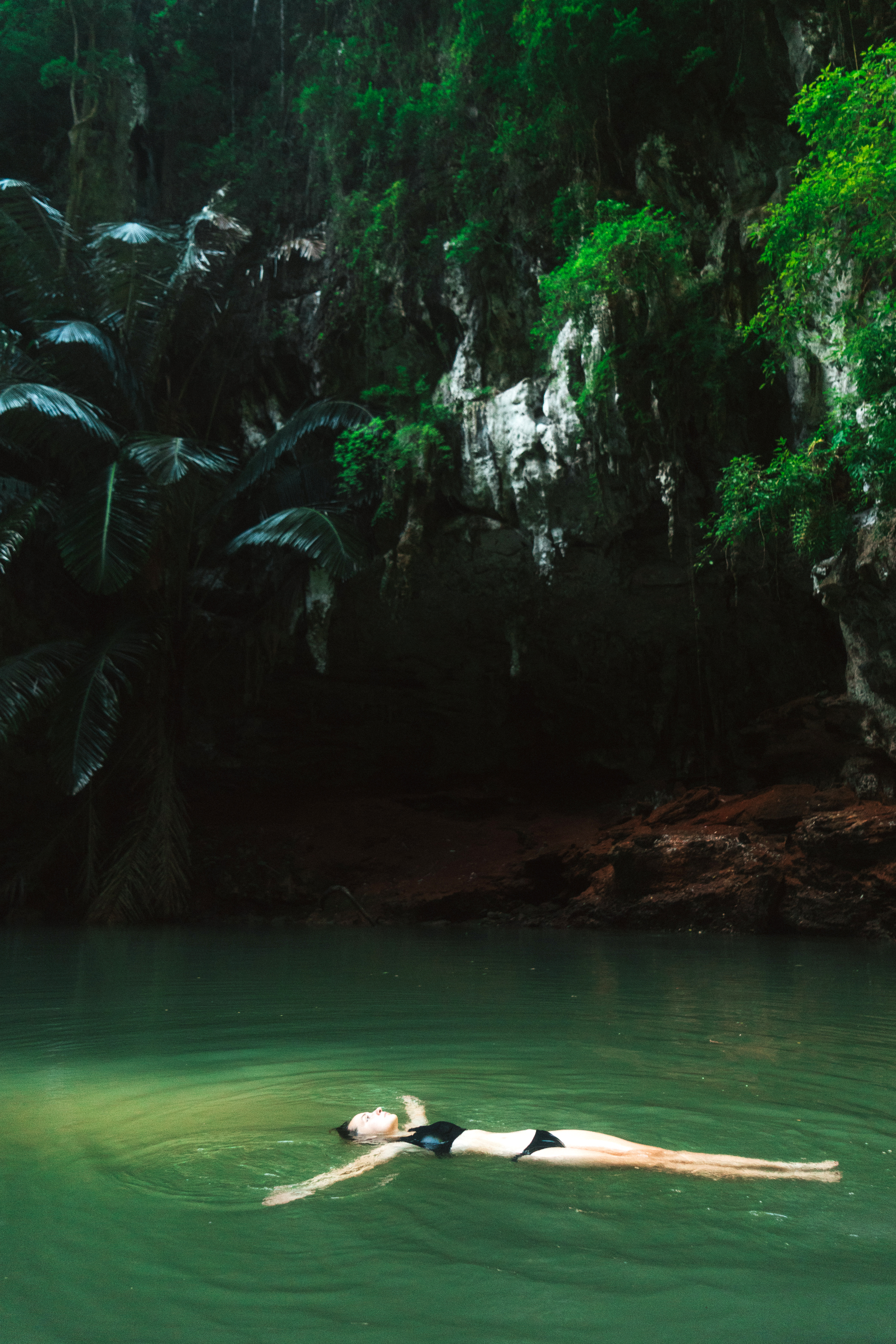 Swimmer glides through emerald water inside a cave lagoon, with sunlight pouring in from an opening above.