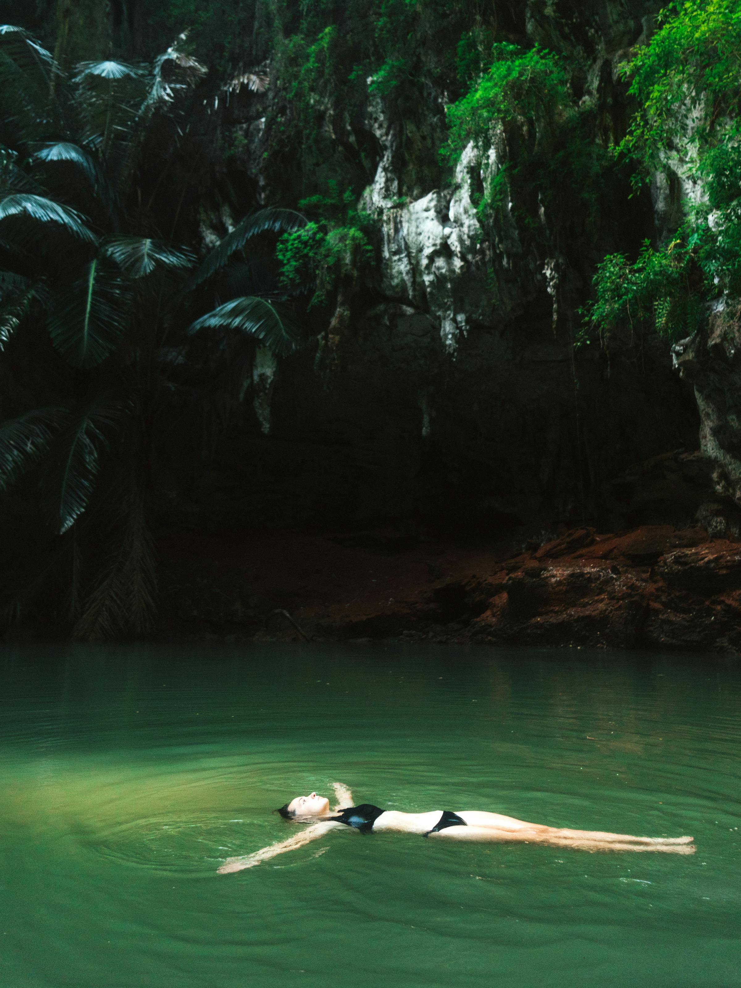 Swimmer glides through emerald water inside a cave lagoon, with sunlight pouring in from an opening above.