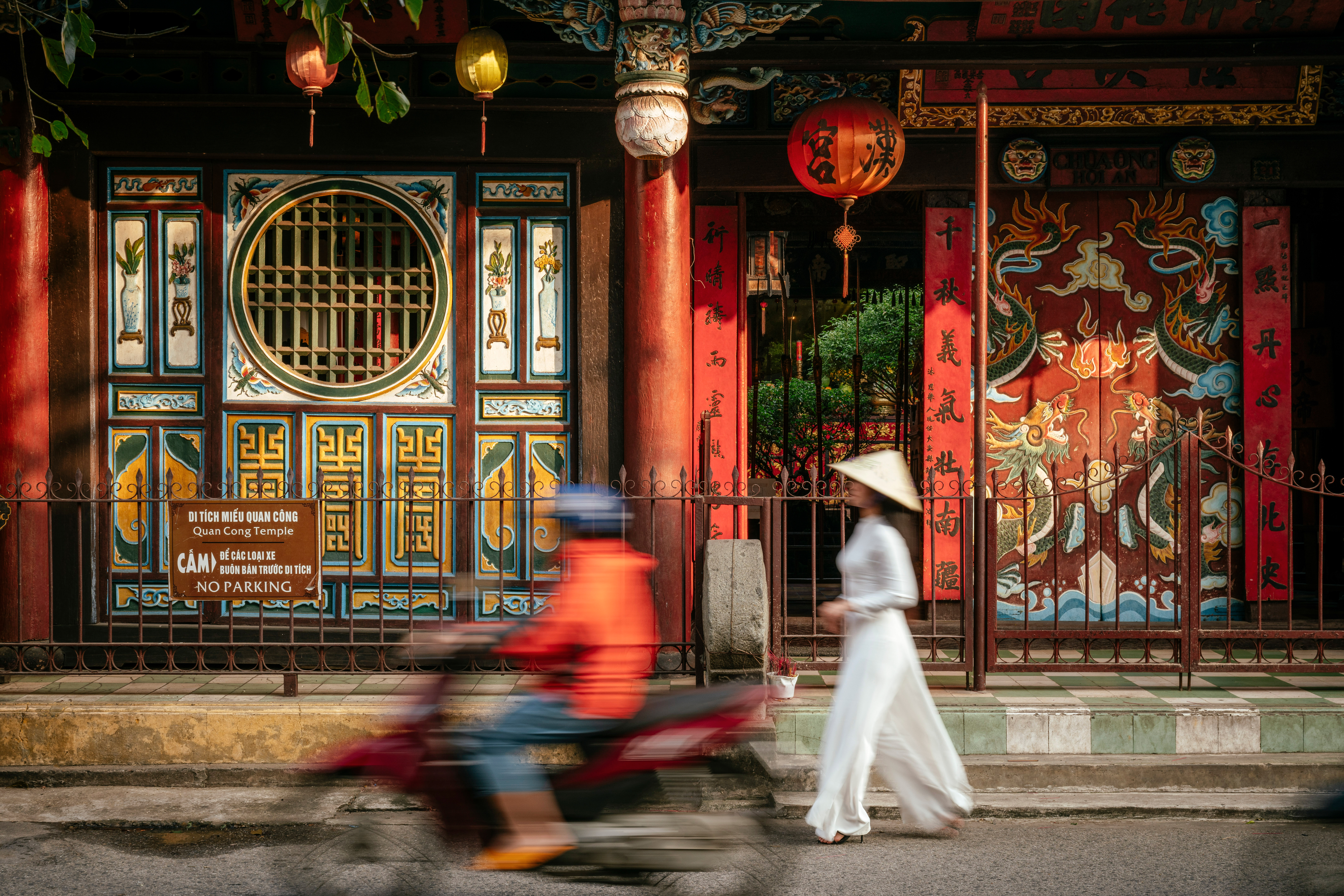 Motorbike blurs past a colorful storefront with hanging lanterns, while a pedestrian walks along the curb.