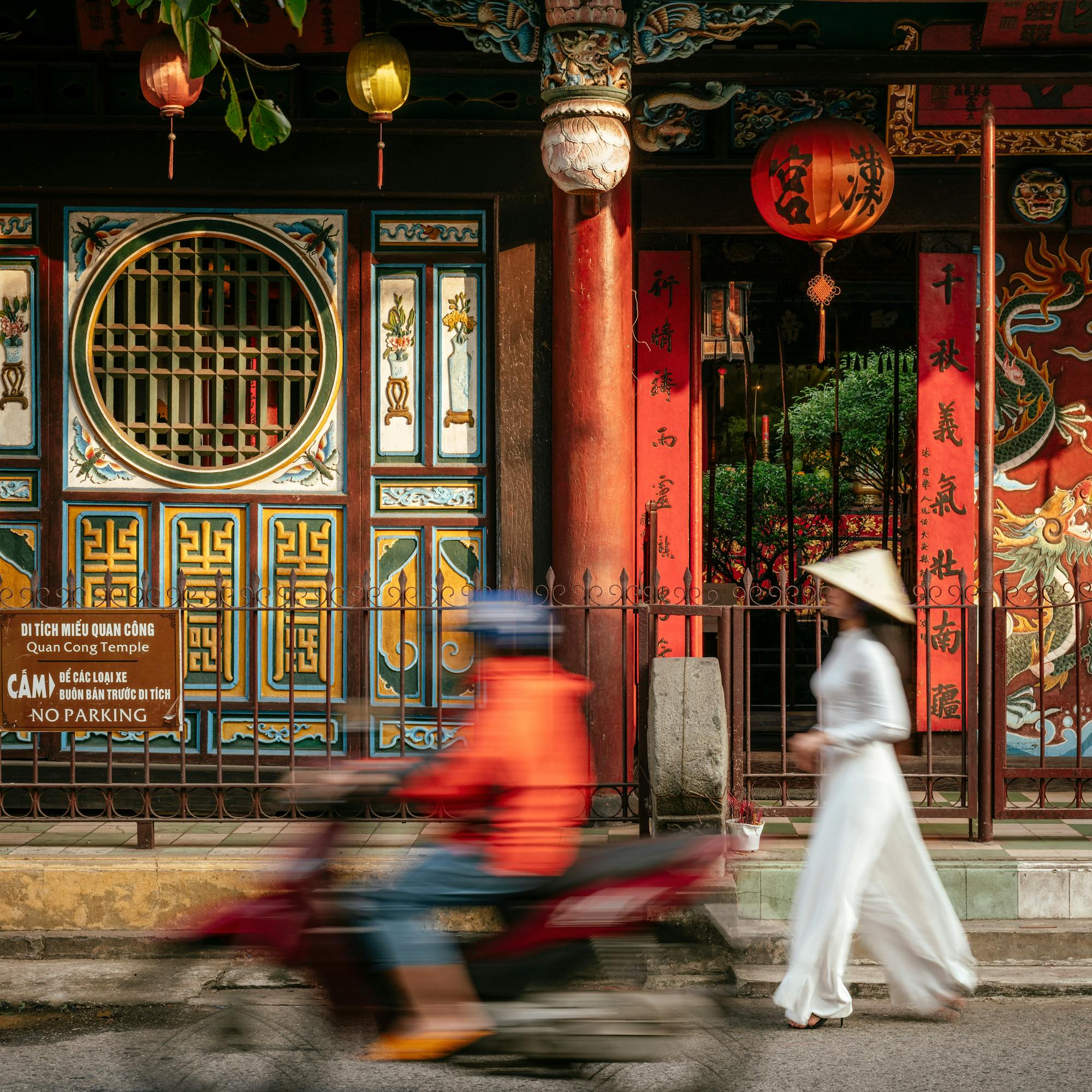 Motorbike blurs past a colorful storefront with hanging lanterns, while a pedestrian walks along the curb.