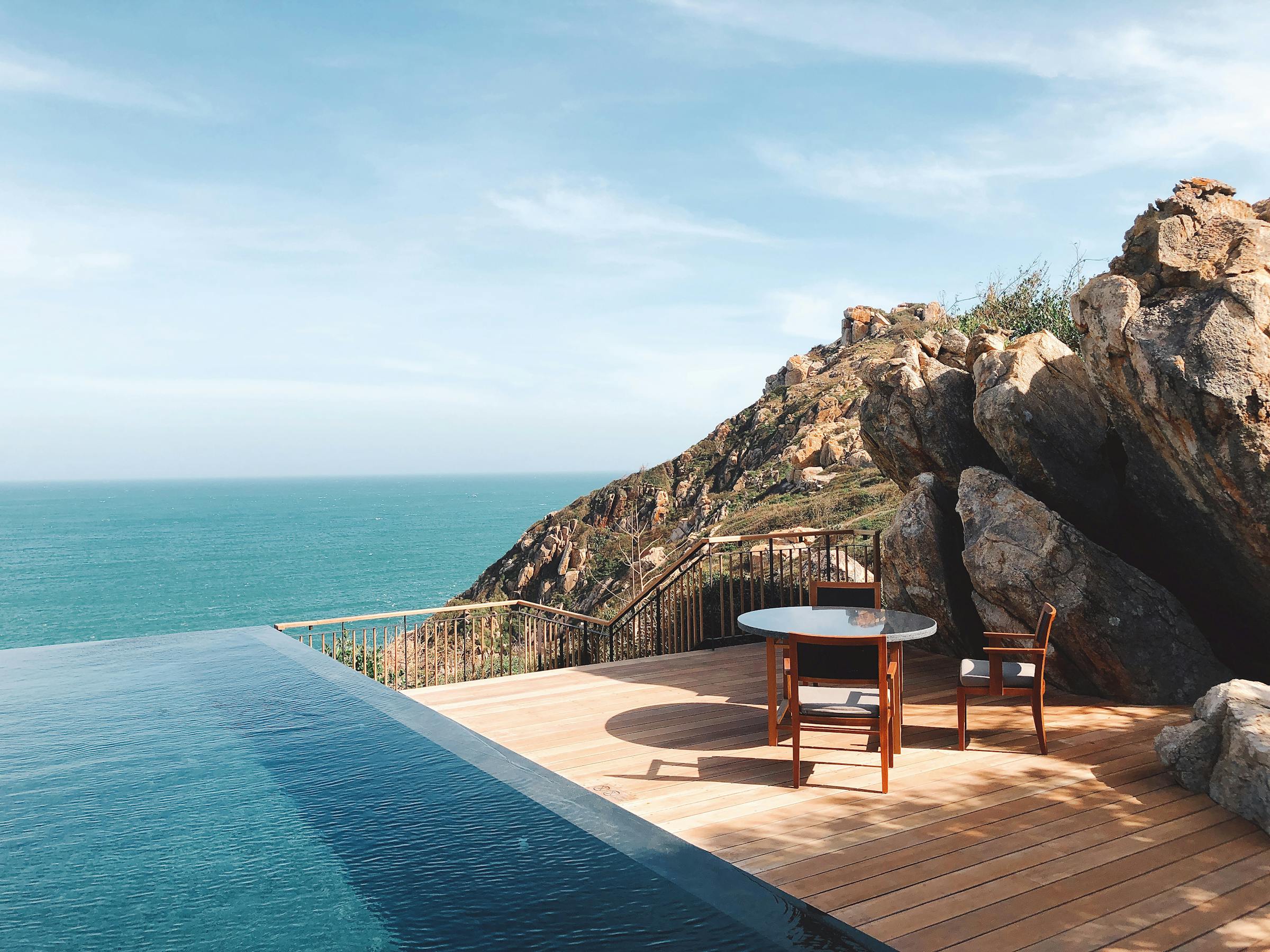 Infinity pool edge meets the horizon, with lounge chairs and a small table set above a deep blue ocean under soft daylight.