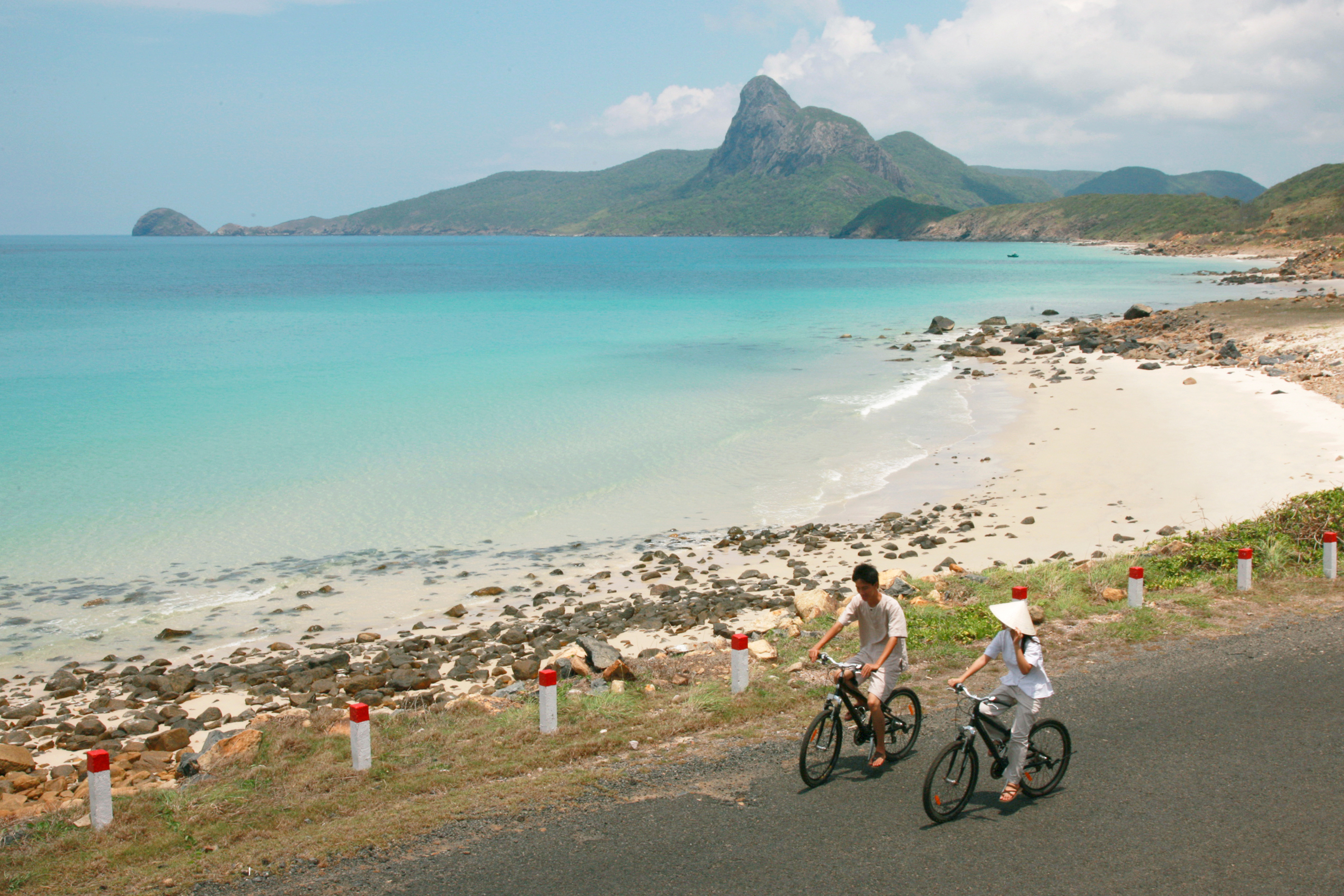 Two bicycles ride along a coastal road beside turquoise water, with sandy shoreline and low hills ahead under soft daylight.