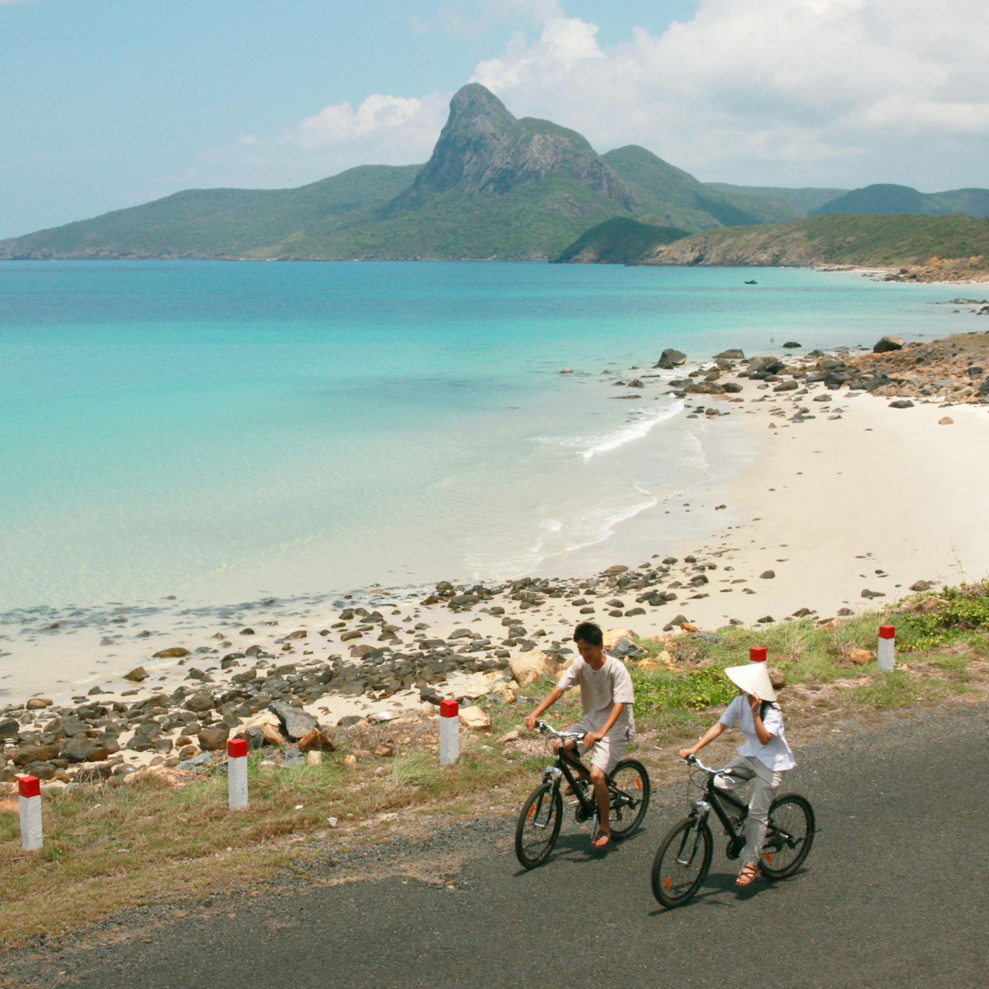 Two bicycles ride along a coastal road beside turquoise water, with sandy shoreline and low hills ahead under soft daylight.