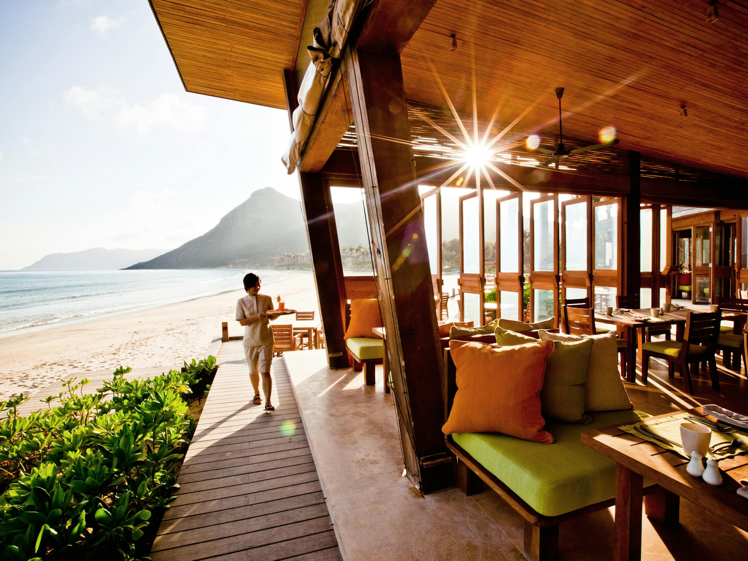 Open-air restaurant deck faces the sea, with wooden tables, chairs, and a walkway leading to the beach under soft daylight.