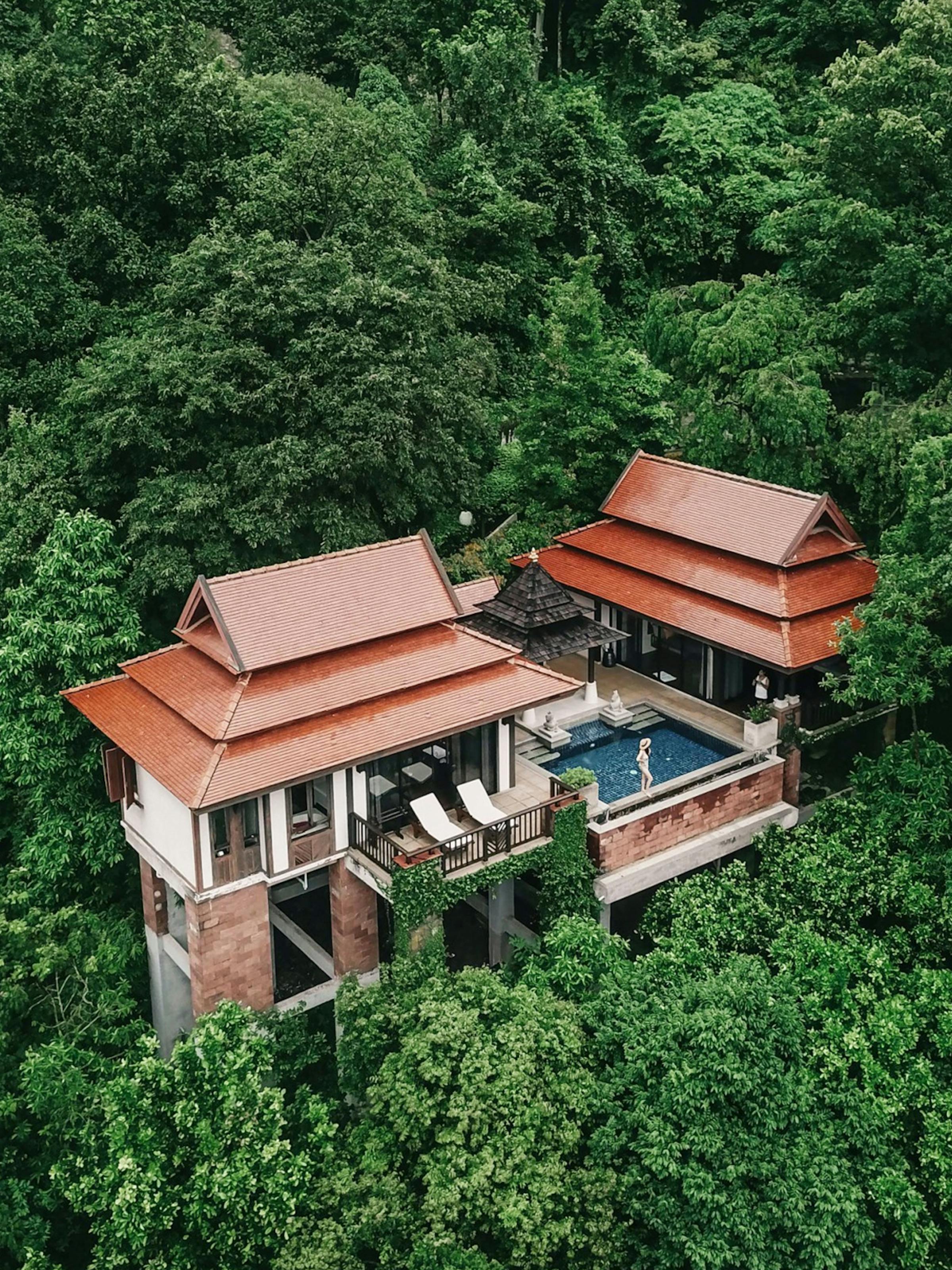 Thatched-roof villa and pool sit above a hillside, with forested slopes under soft daylight.
