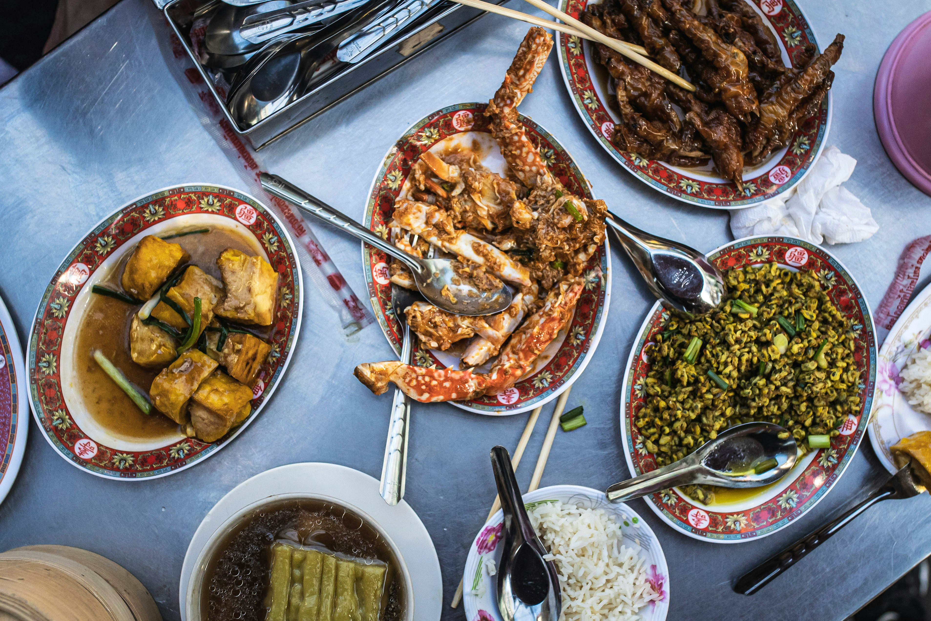 Bowls and plates of street food cover a table, with sauces, herbs, and grilled dishes arranged in rows under soft daylight.