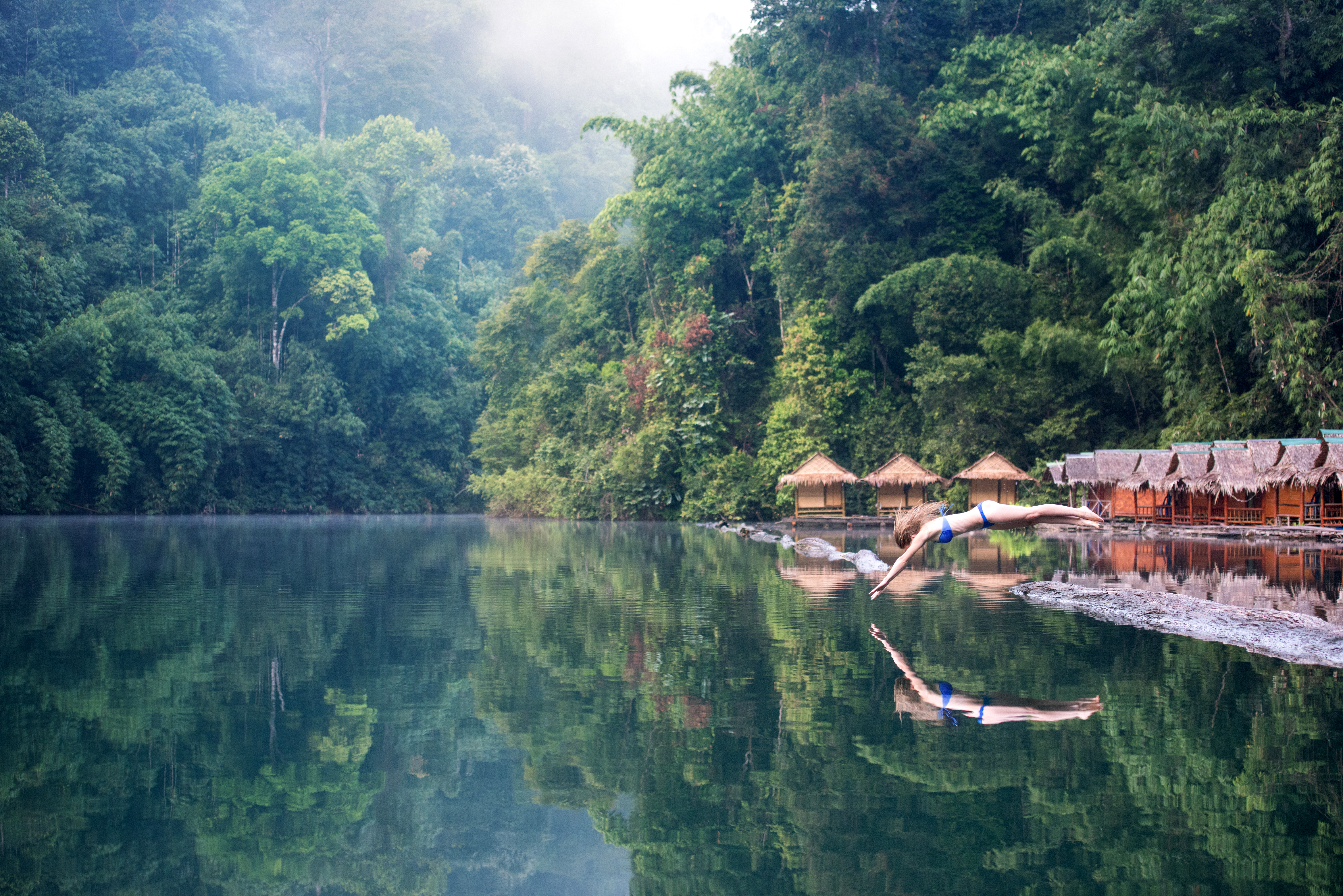 Person dives into an emerald lake surrounded by steep forested cliffs, with mist over the water under soft daylight.