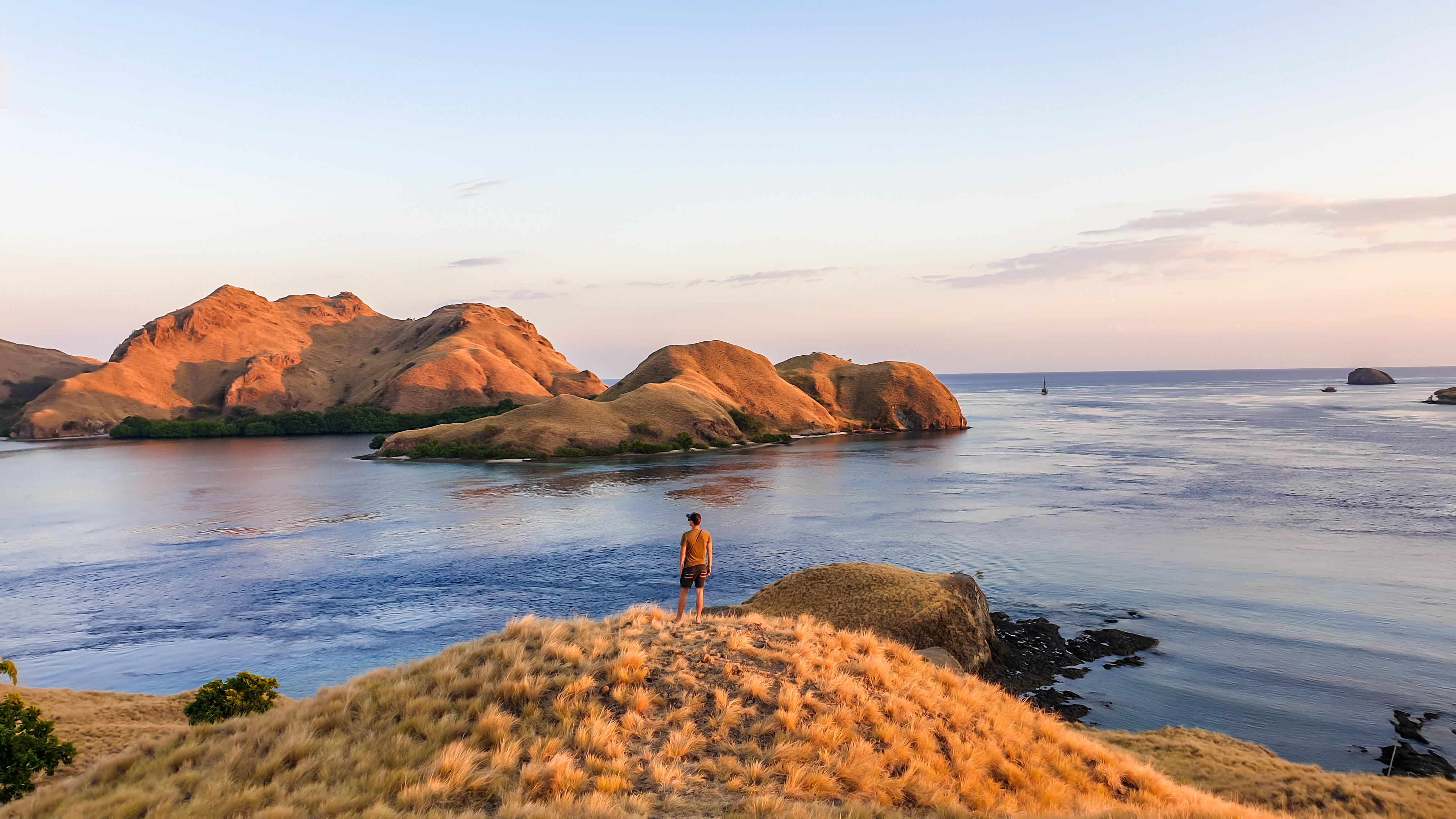 Person stands on a rocky headland overlooking a wide bay, with cliffs and blue water below the horizon under warm morning light.