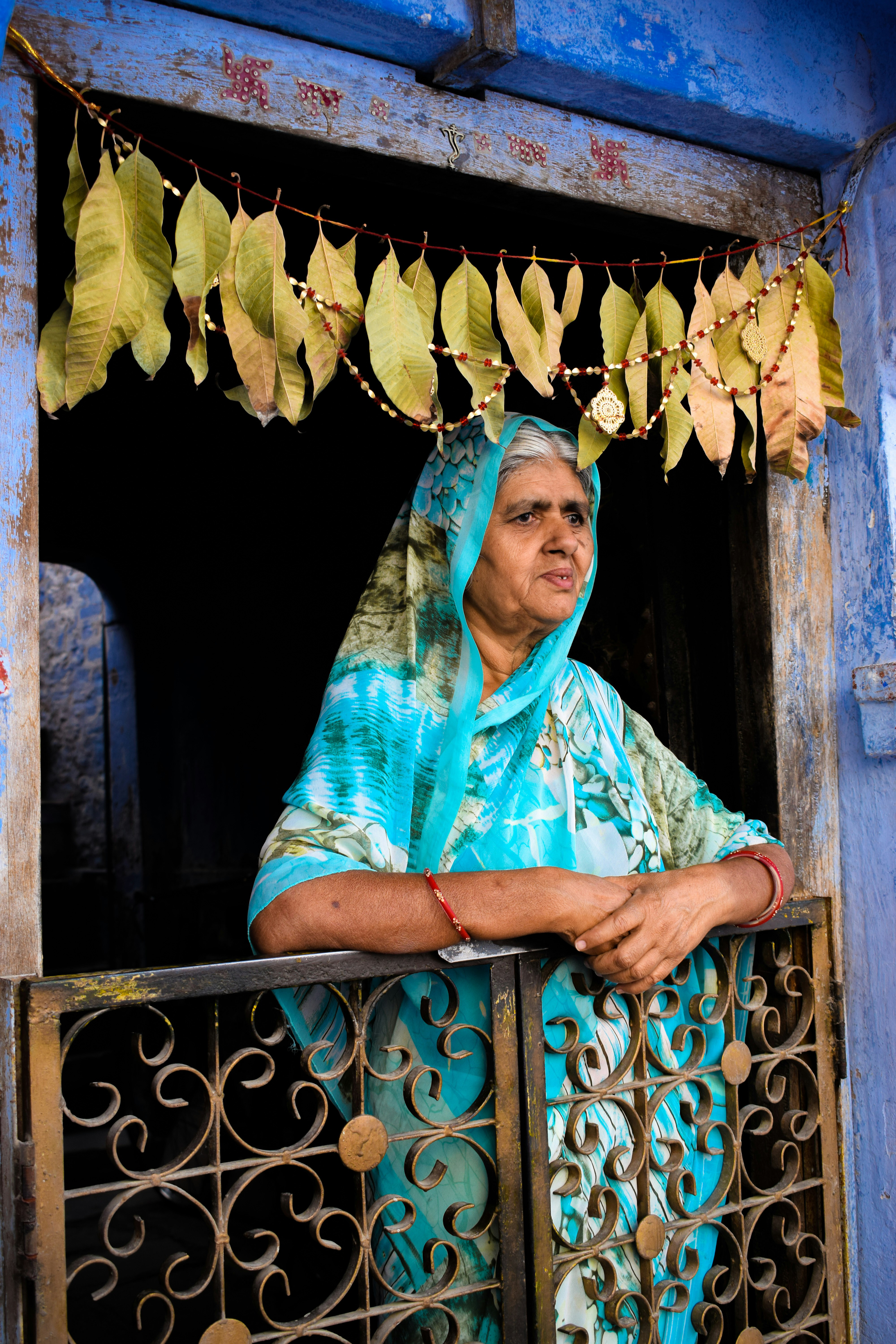 A woman in a blue sari leans on a balcony in a blue-painted doorway, framed by hanging marigold garlands.