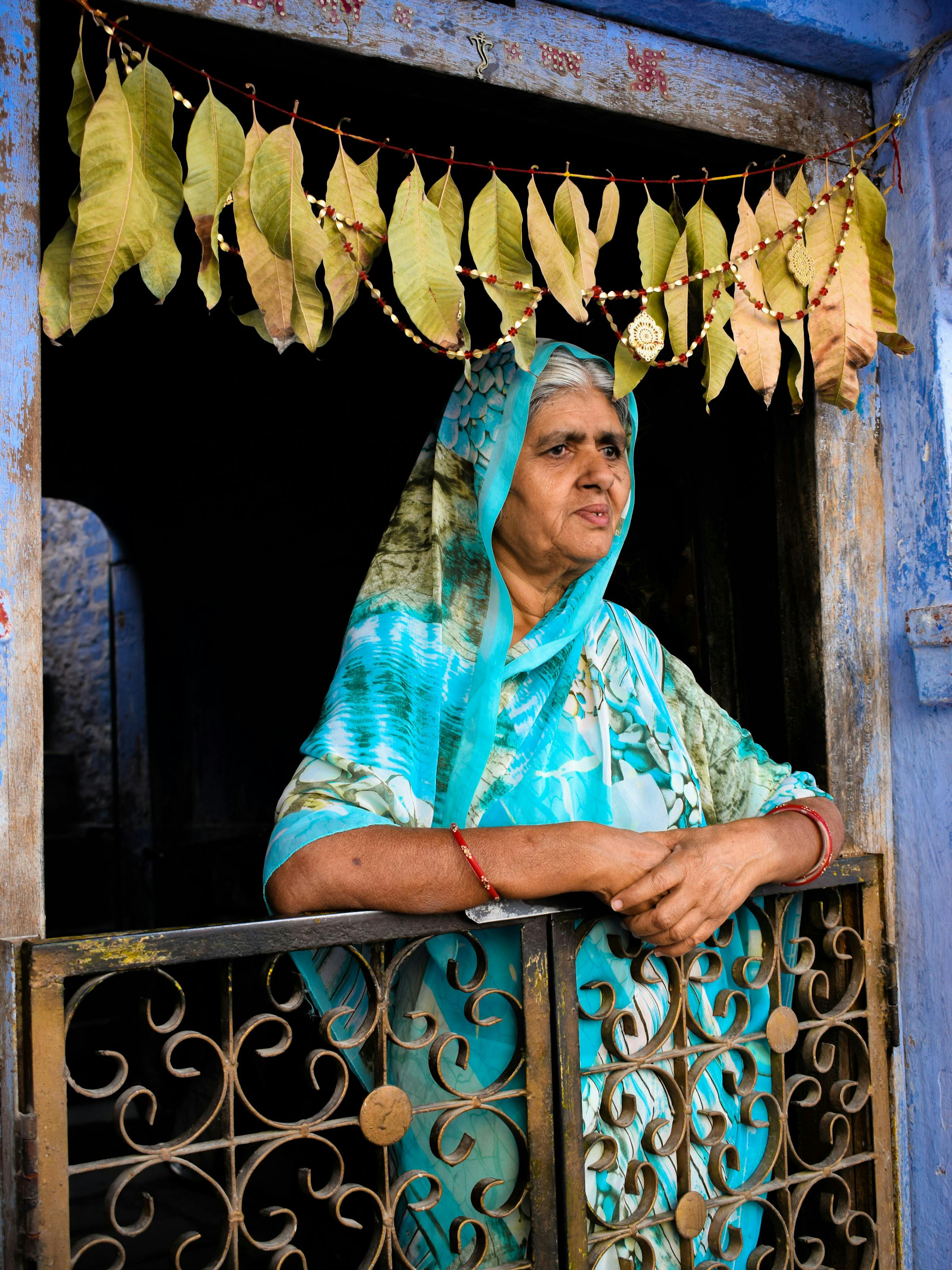 A woman in a blue sari leans on a balcony in a blue-painted doorway, framed by hanging marigold garlands.