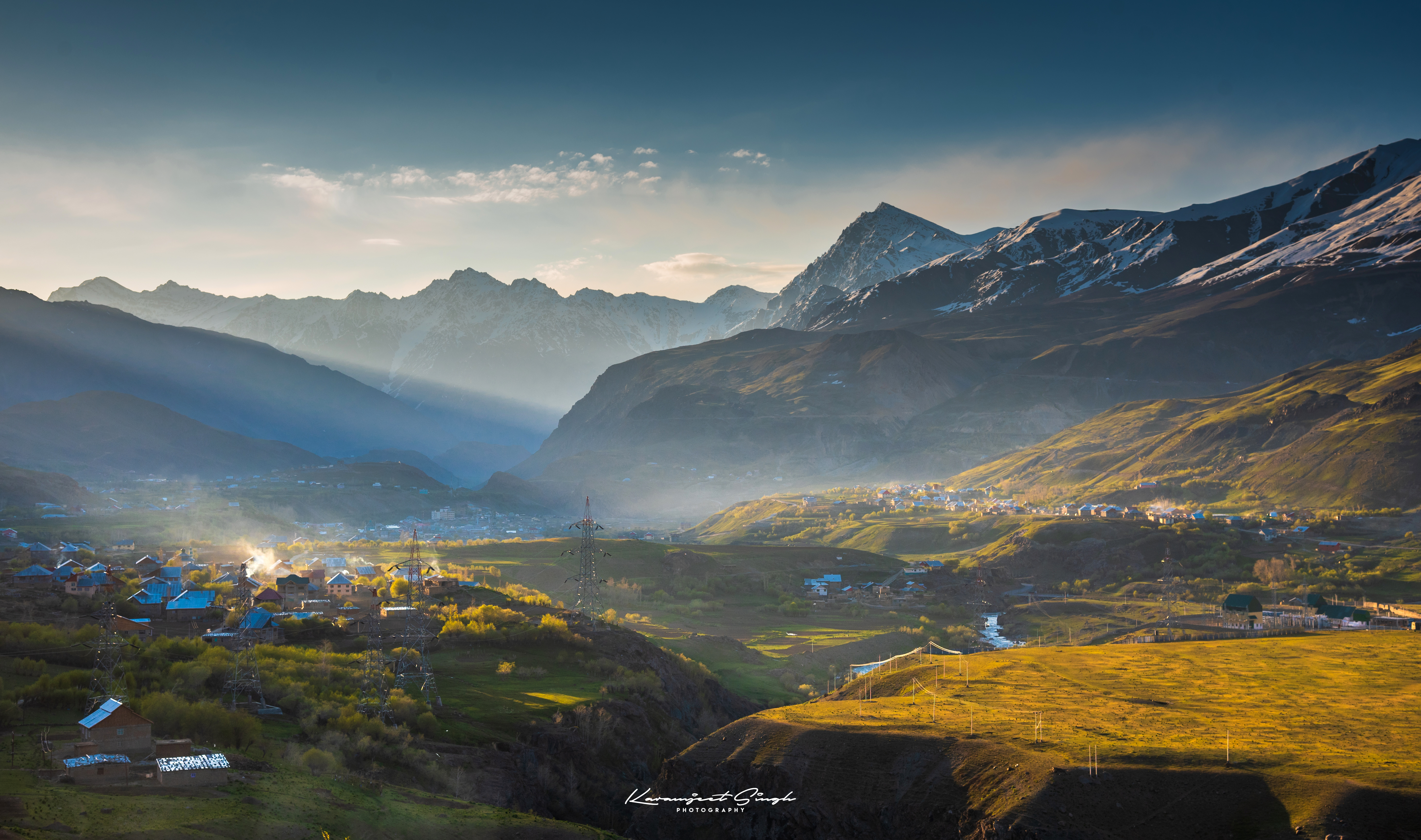 Sunlit green fields and scattered homes fill a mountain valley, with layered peaks fading into blue distance.