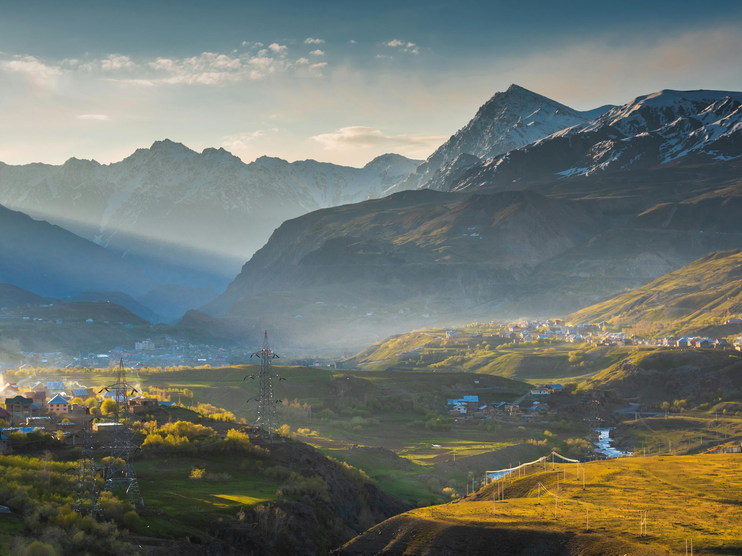 Sunlit green fields and scattered homes fill a mountain valley, with layered peaks fading into blue distance.