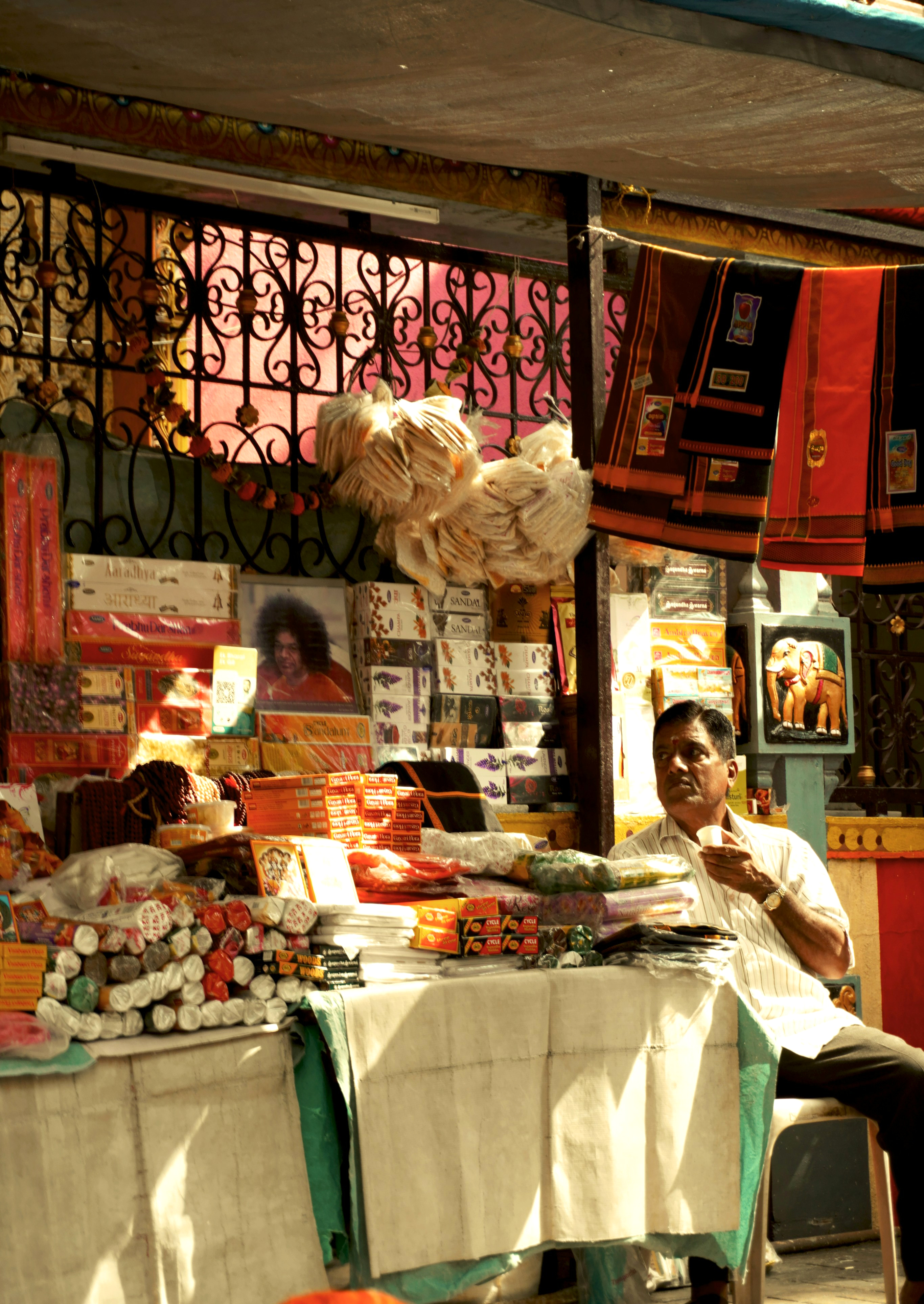A colorful market stall displays textiles and souvenirs, with sunlight slanting across hanging fabrics and shelves.
