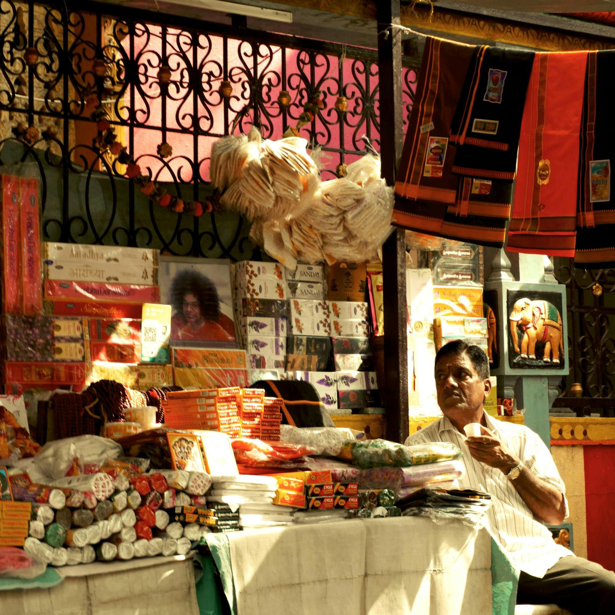 A colorful market stall displays textiles and souvenirs, with sunlight slanting across hanging fabrics and shelves.