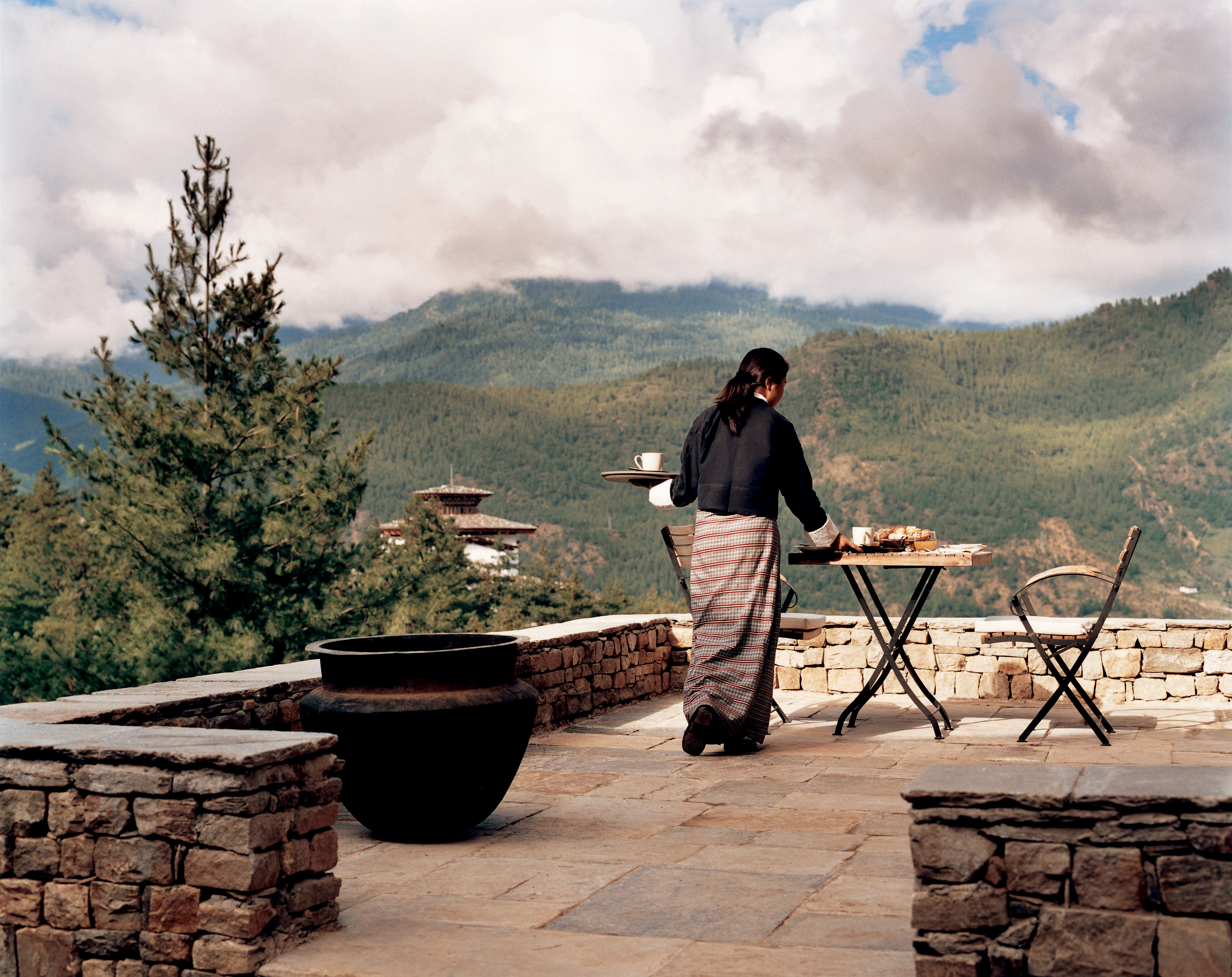 A person stands by a terrace table set for dining, overlooking rolling hills and a valley under soft overcast light.