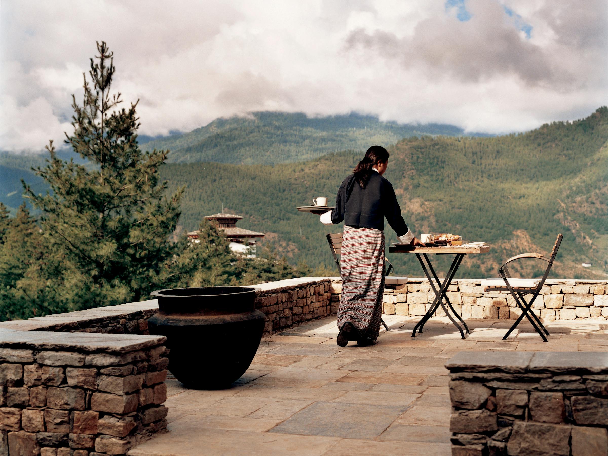 A person stands by a terrace table set for dining, overlooking rolling hills and a valley under soft overcast light.