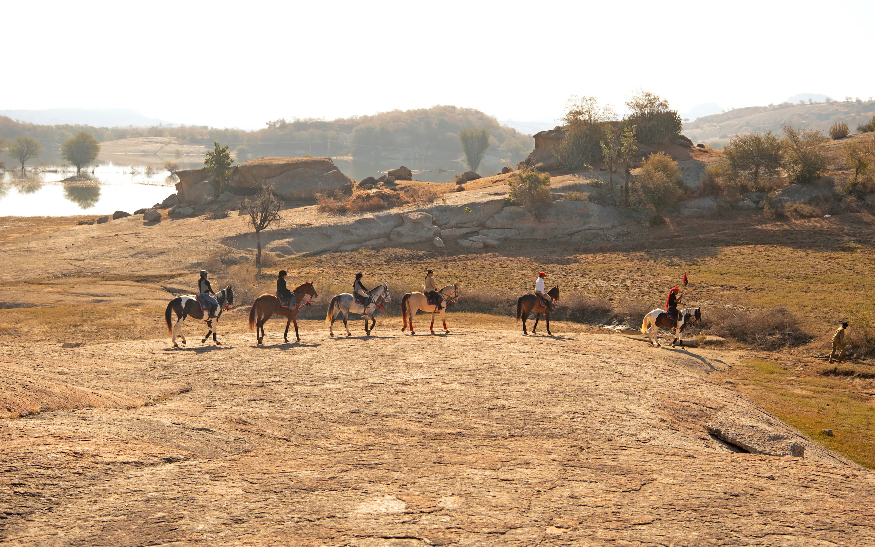 A line of horses with riders crosses a rocky desert plain, with long shadows and a hazy horizon in the distance.
