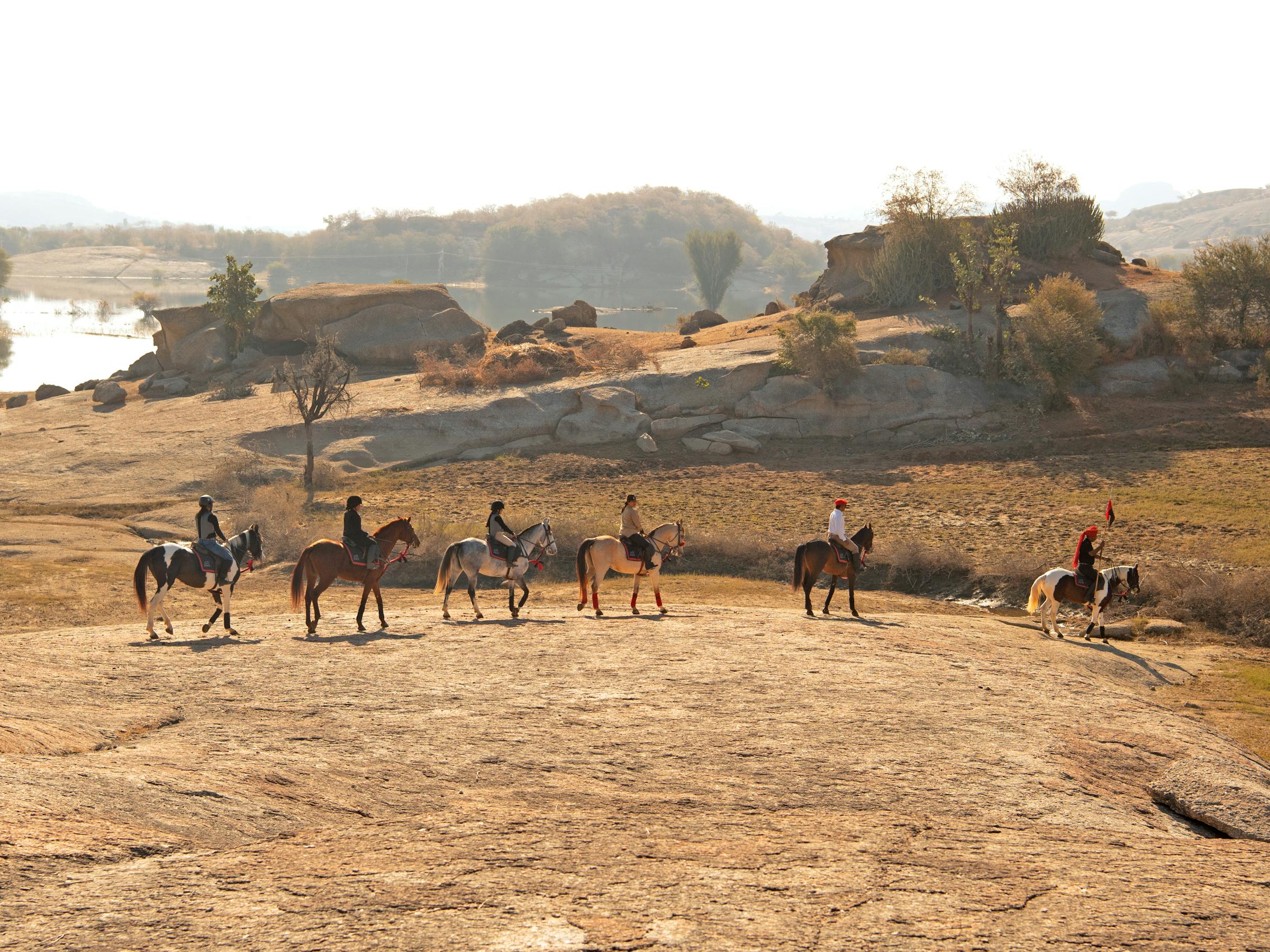 A line of horses with riders crosses a rocky desert plain, with long shadows and a hazy horizon in the distance.