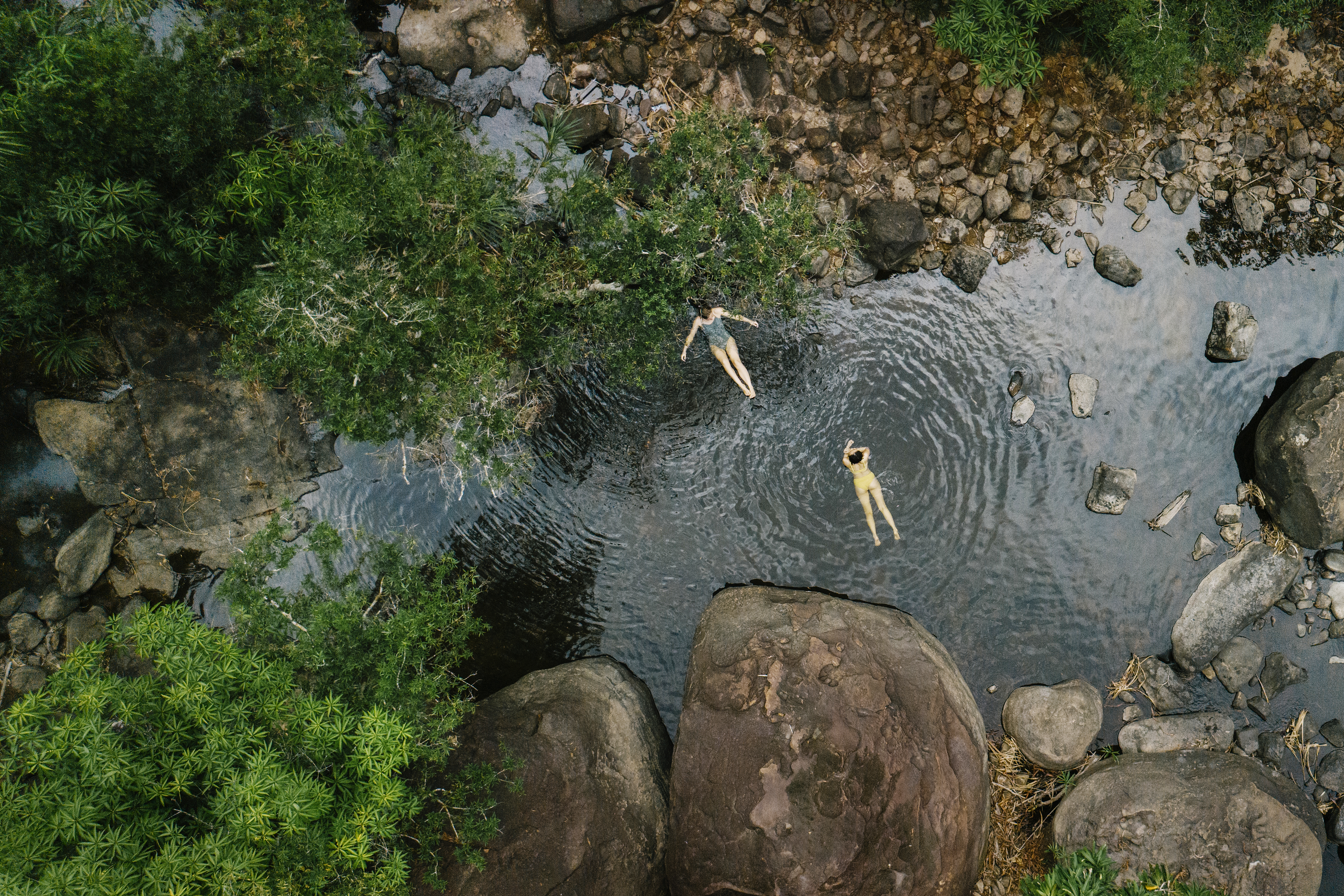 People swim in a rocky river pool, seen from above, with boulders and clear water winding through trees.