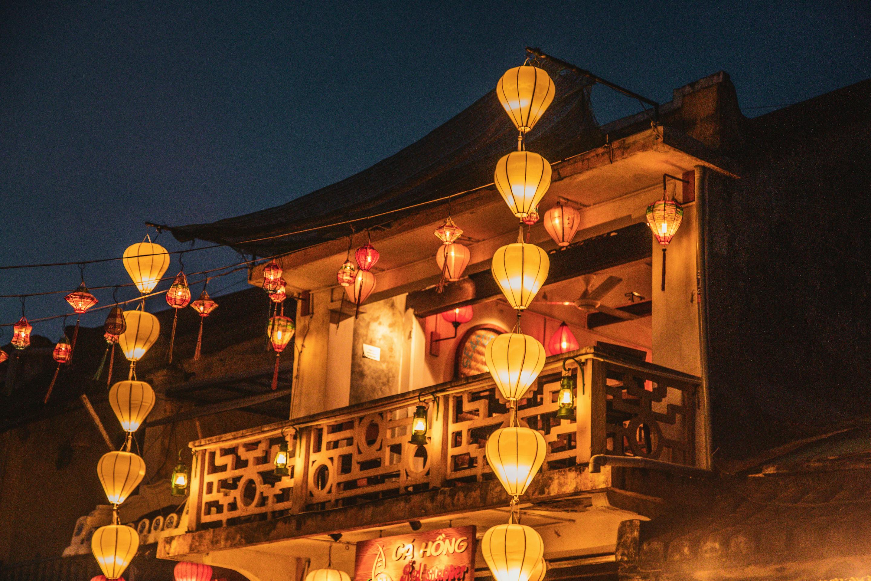 Lantern-lit balcony glows at night, with warm lights hanging from a wooden facade above a dark street under soft daylight.