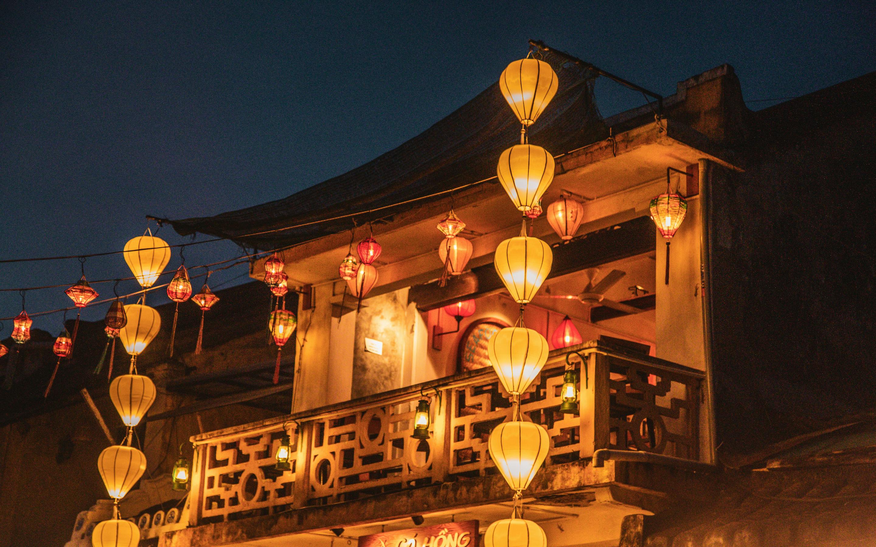 Lantern-lit balcony glows at night, with warm lights hanging from a wooden facade above a dark street under soft daylight.