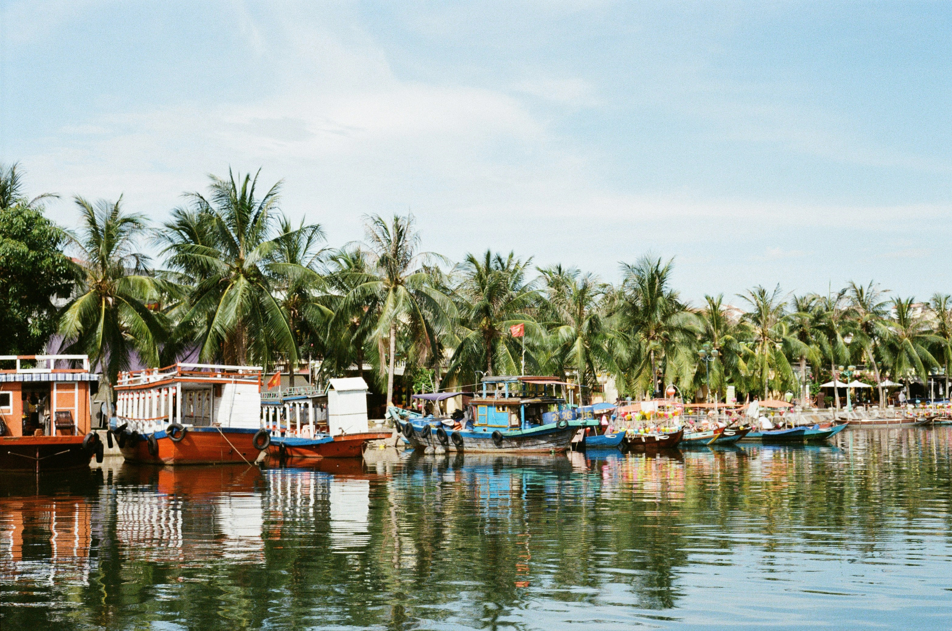 Boats rest along a calm river bordered by palm trees, with reflections on the water under a pale sky under soft daylight.
