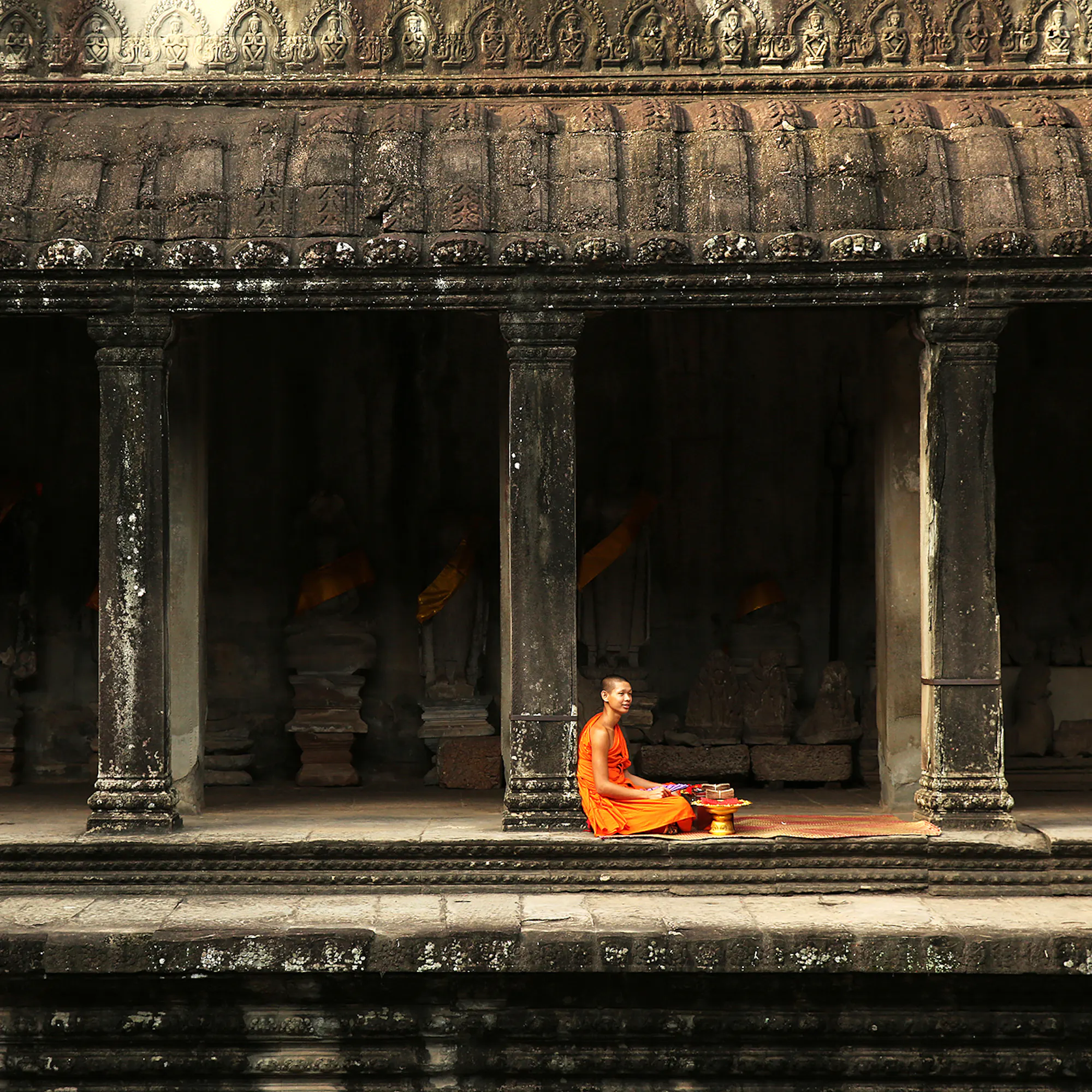 Person in orange robe sits in a stone temple corridor, with carved columns and deep shadows behind under soft daylight.