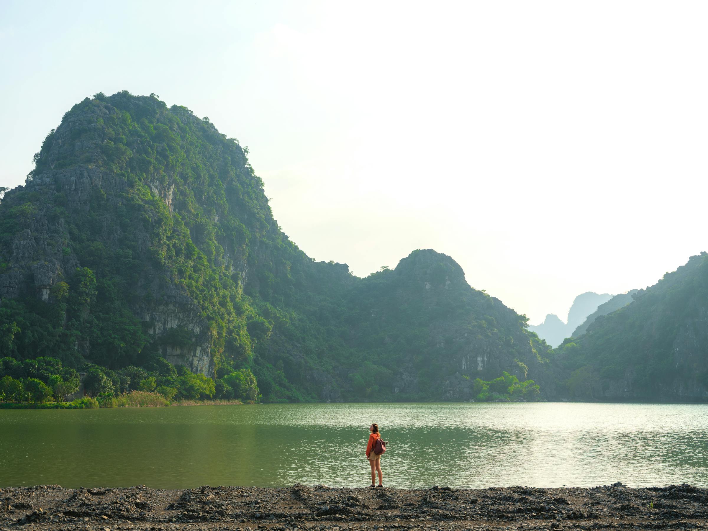 Person stands on a sandy shore facing limestone karsts rising from calm water beneath a hazy sky under soft daylight.